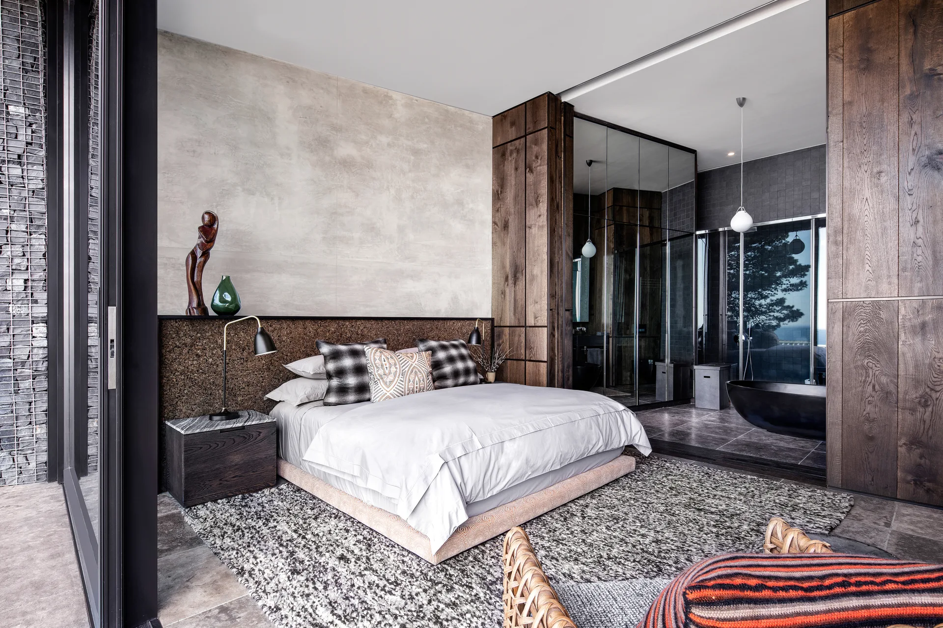 A modern bedroom features a low bed with a cork headboard panel, wood accent walls, and an open en-suite bathroom with a black freestanding tub.