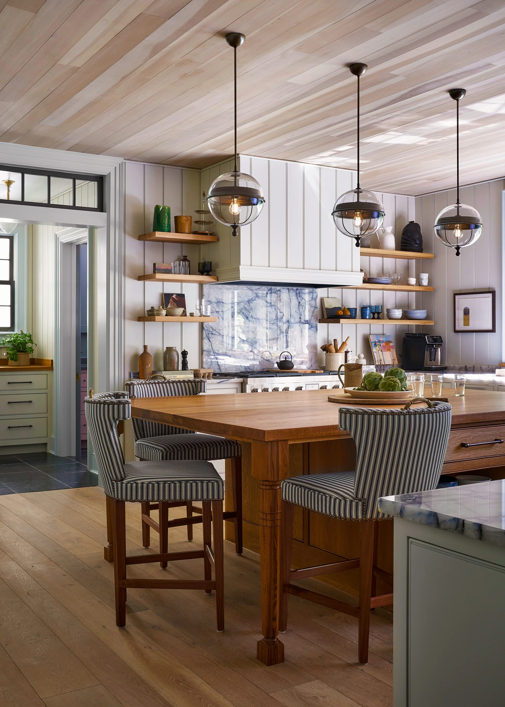 A farmhouse kitchen features a light wood plank ceiling, a central wood island with striped stools, and a blue veined marble backsplash.