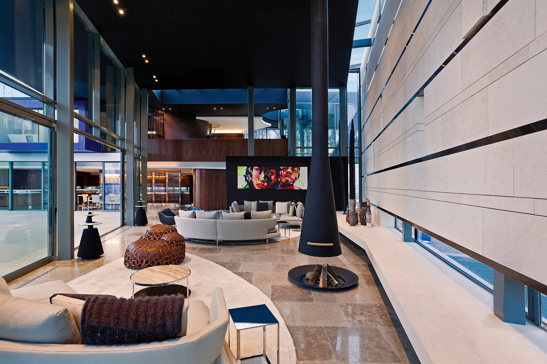 A spacious modern living room with curved beige sofas, a black suspended cone fireplace, and a long horizontal stone paneled wall.