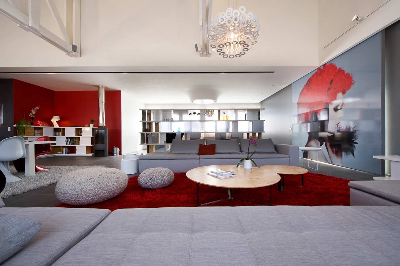 A modern open-plan living room features gray modular sofas, a vibrant red shag rug, round wooden coffee tables, and a large red wall mural.