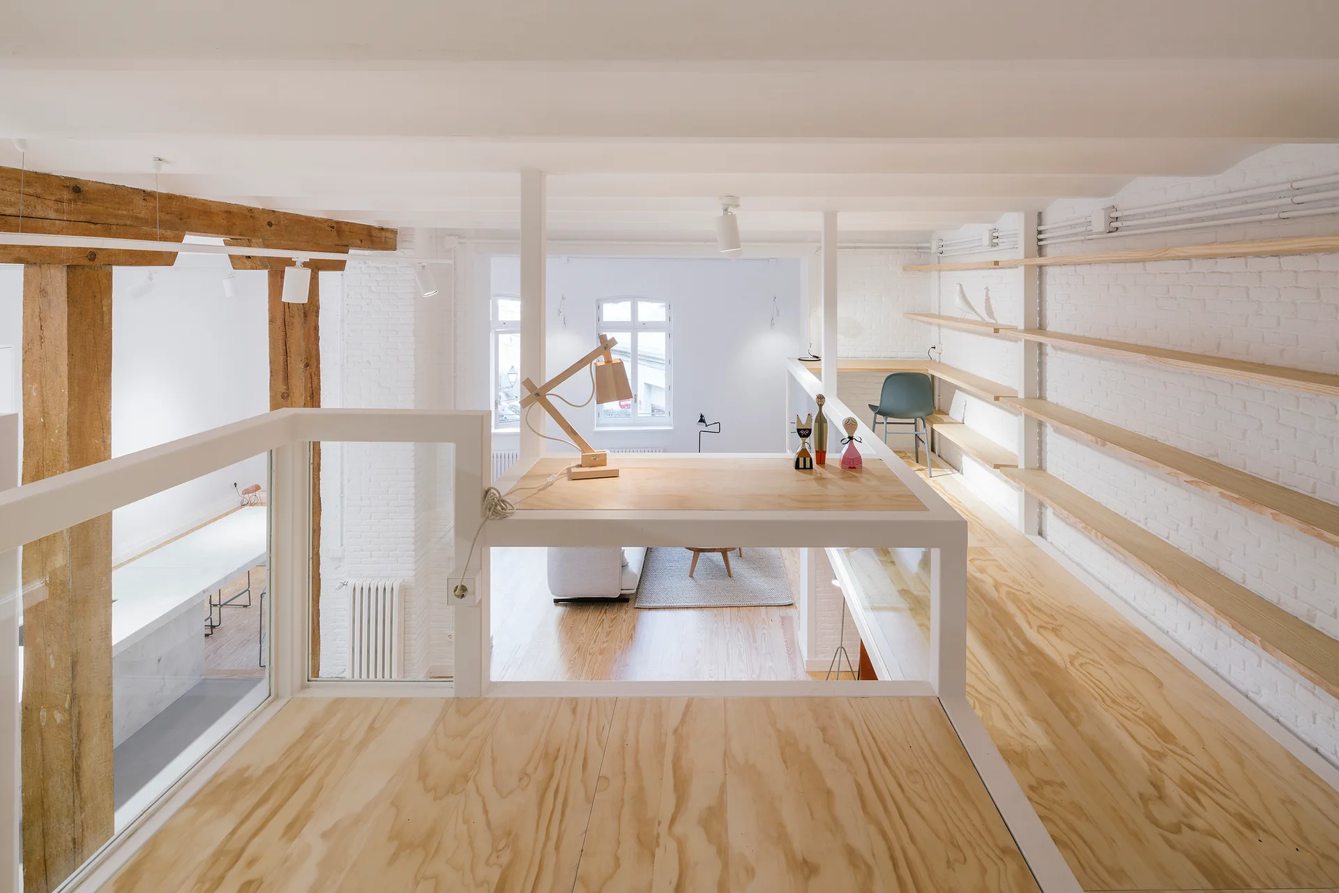 A loft office space features plywood floors, a built-in wooden desk overlooking a lower level, and long wood shelves on a white brick wall.