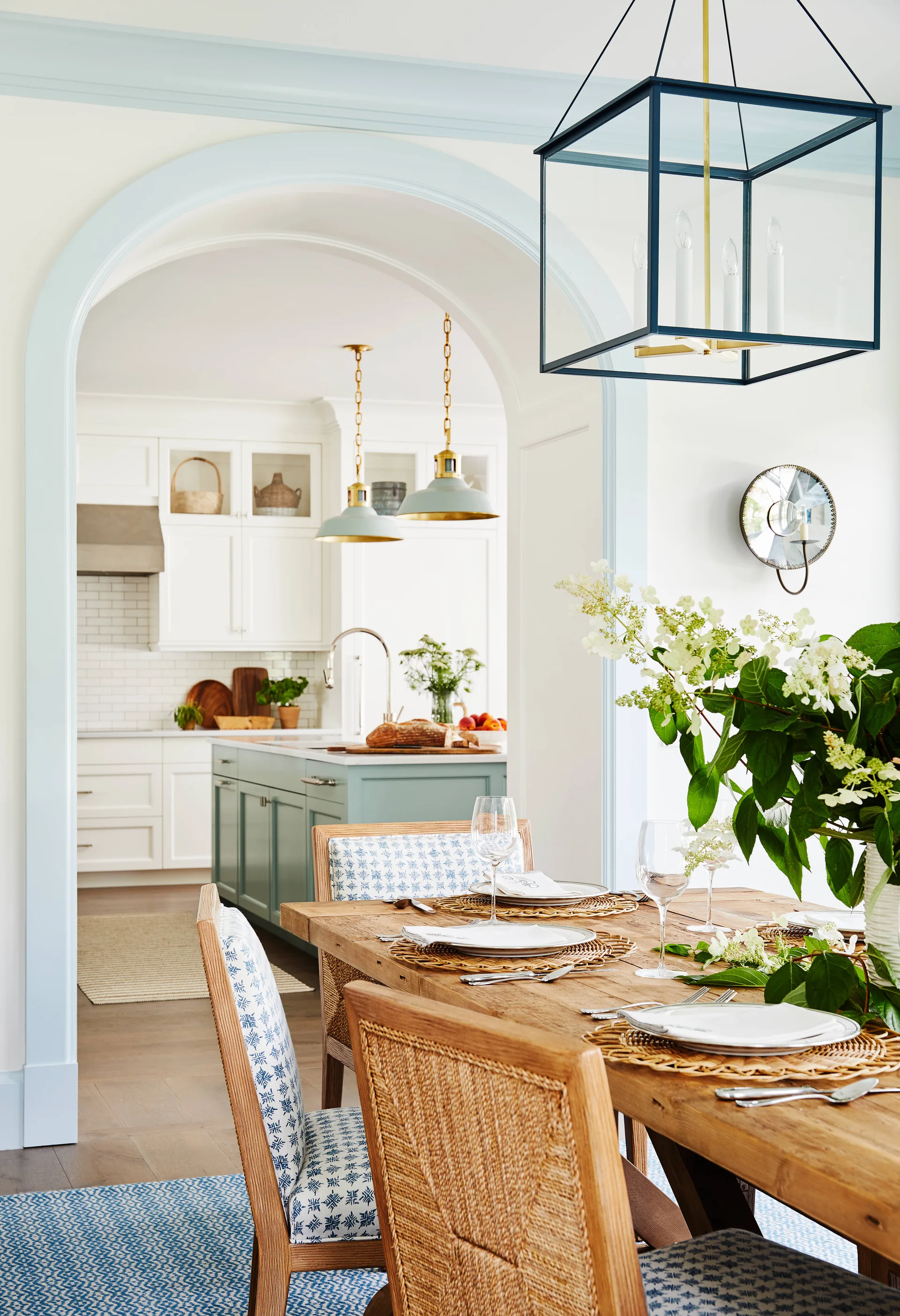 A wooden dining table set with woven placemats sits before a pale blue arched opening that looks into a white kitchen with a matching blue island.