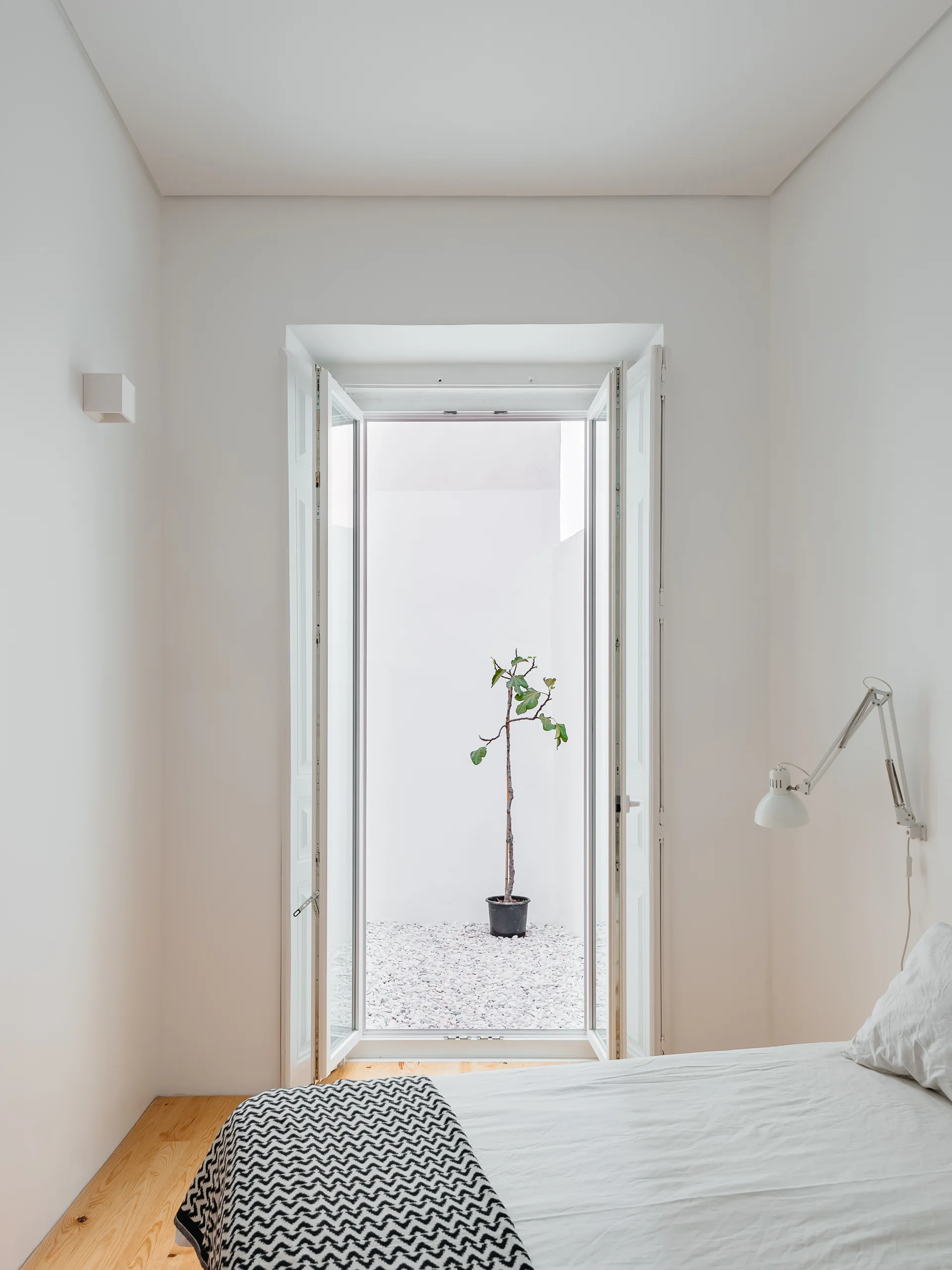 A minimalist white bedroom with light wood floors and tall french doors opening to a private gravel courtyard with a single potted plant.
