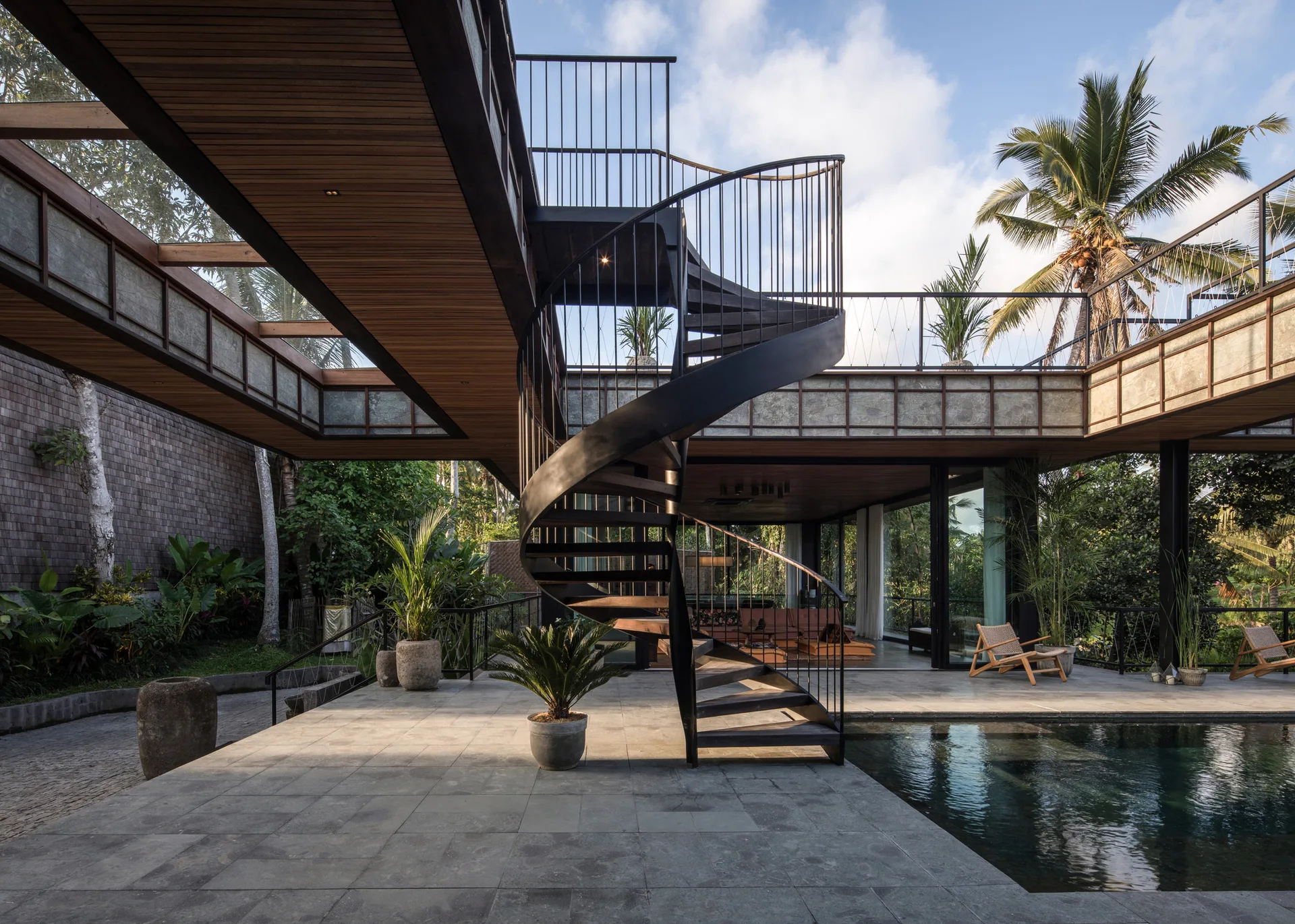 A modern outdoor courtyard with a central black metal spiral staircase, a stone tile patio next to a swimming pool, and an overhead wooden walkway.