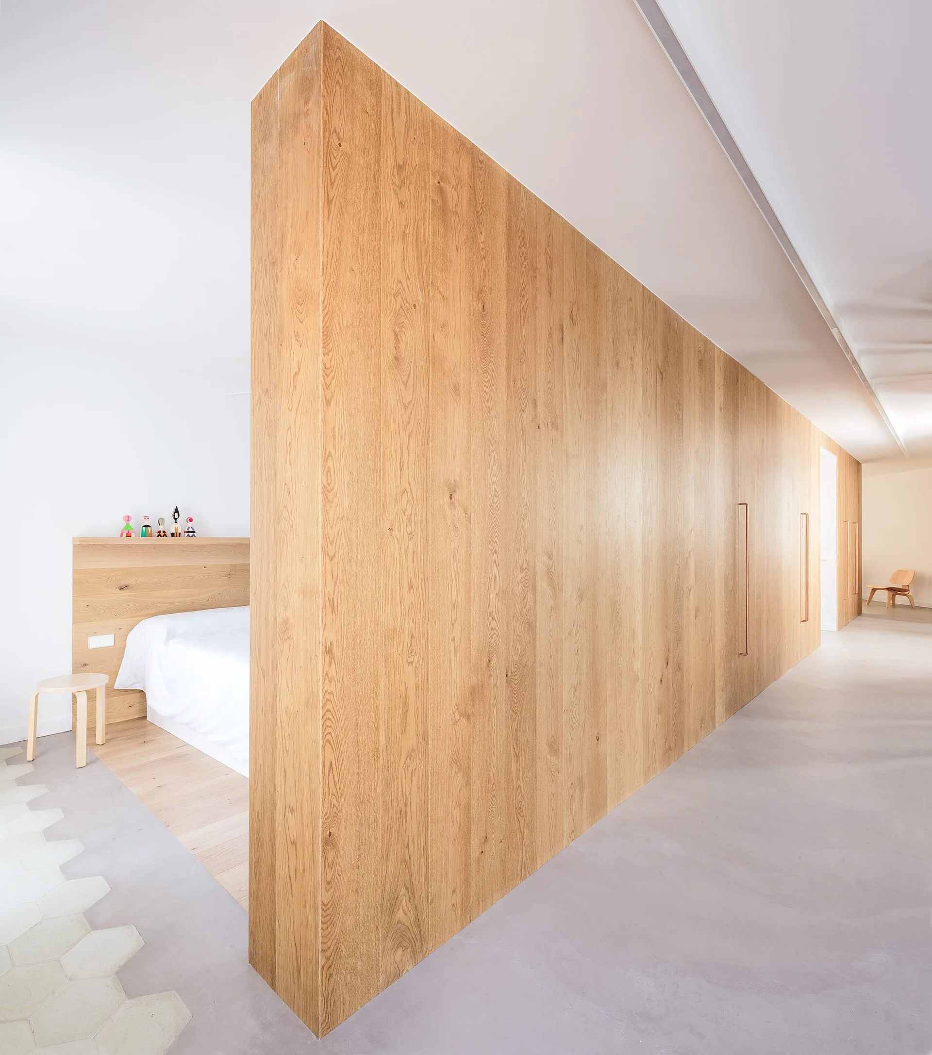 A minimalist bedroom with a bed and light wood headboard separated from a concrete-floored hallway by a large wood partition wall.