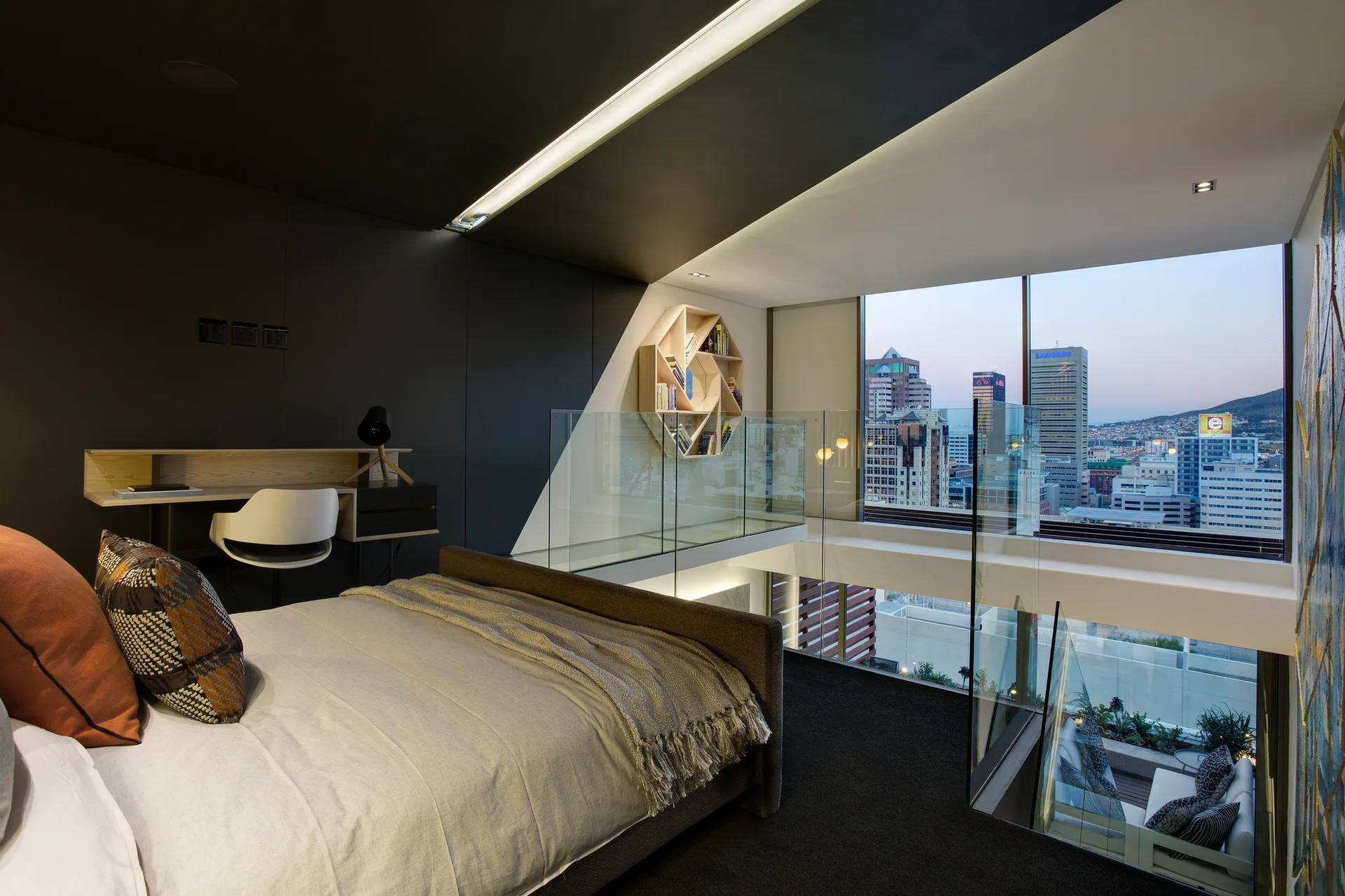 A modern mezzanine bedroom features an upholstered bed, a built-in wooden desk, a hexagonal wall shelf, and glass railings overlooking a city view.