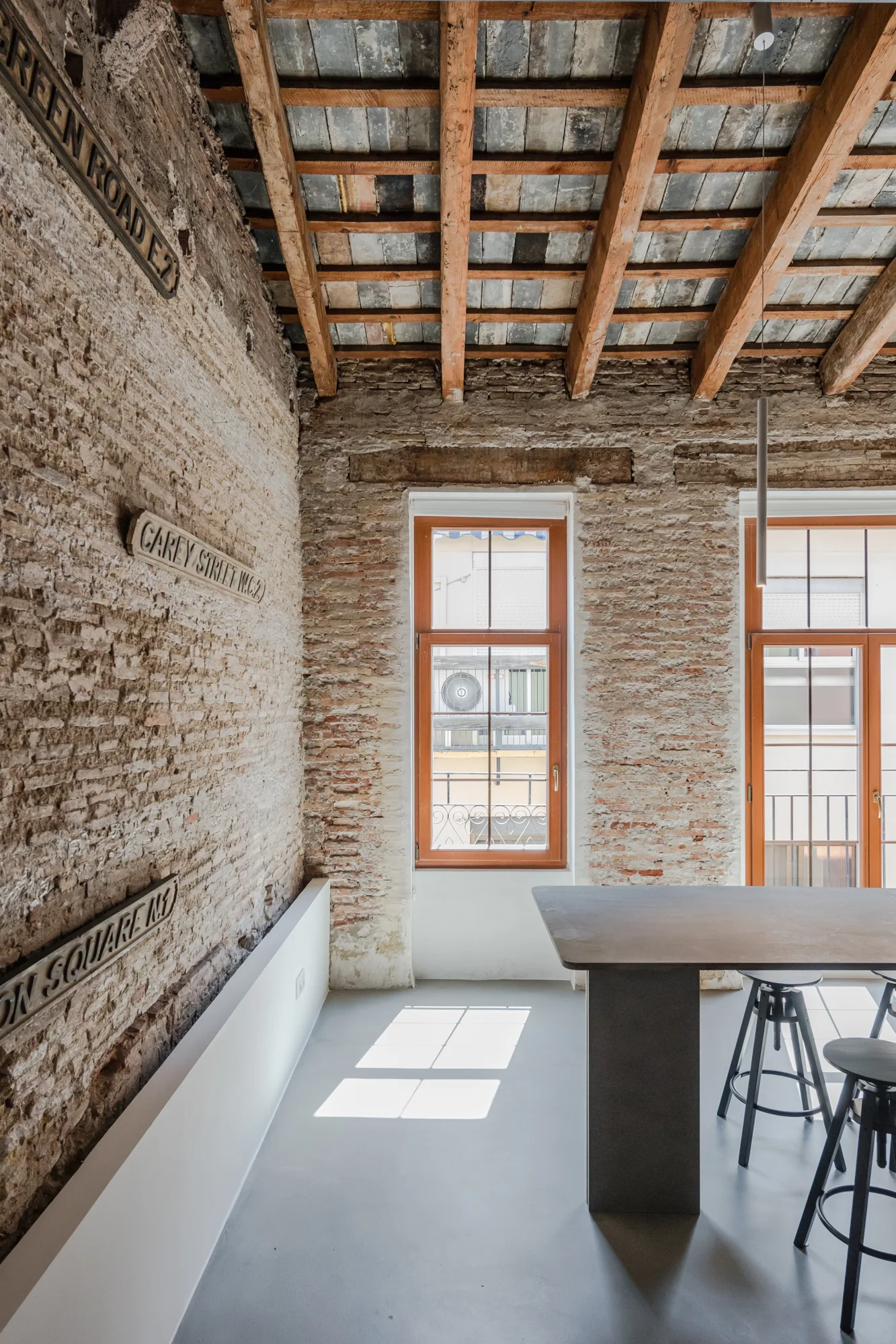 An industrial dining space with exposed brick walls, a wooden beam ceiling, a large concrete table, black metal stools, and street signs.