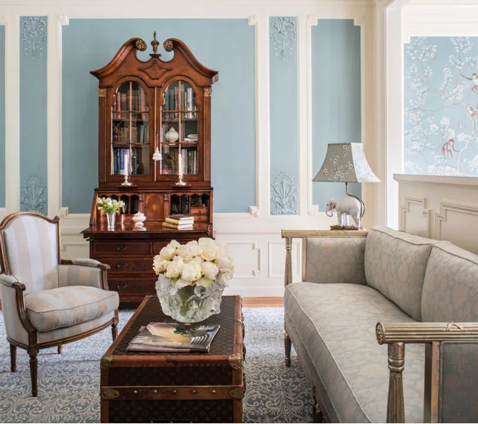 A traditional sitting room with blue paneled walls, an antique wooden secretary desk, a patterned sofa, and a vintage trunk used as a coffee table.