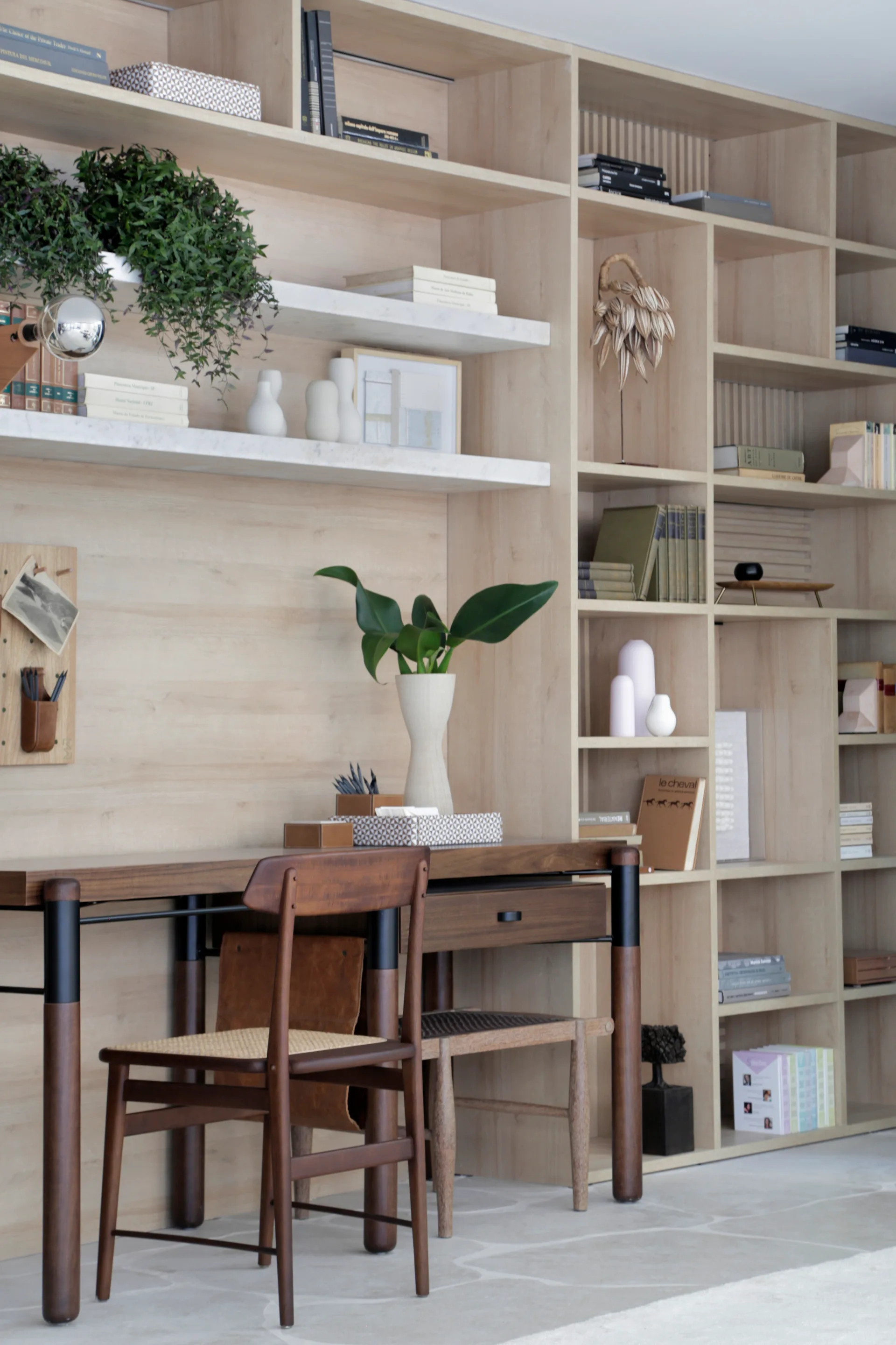 A light wood built-in shelving unit covers the wall behind a dark wood desk, a matching chair with a cane seat, and various books and ceramic vases.