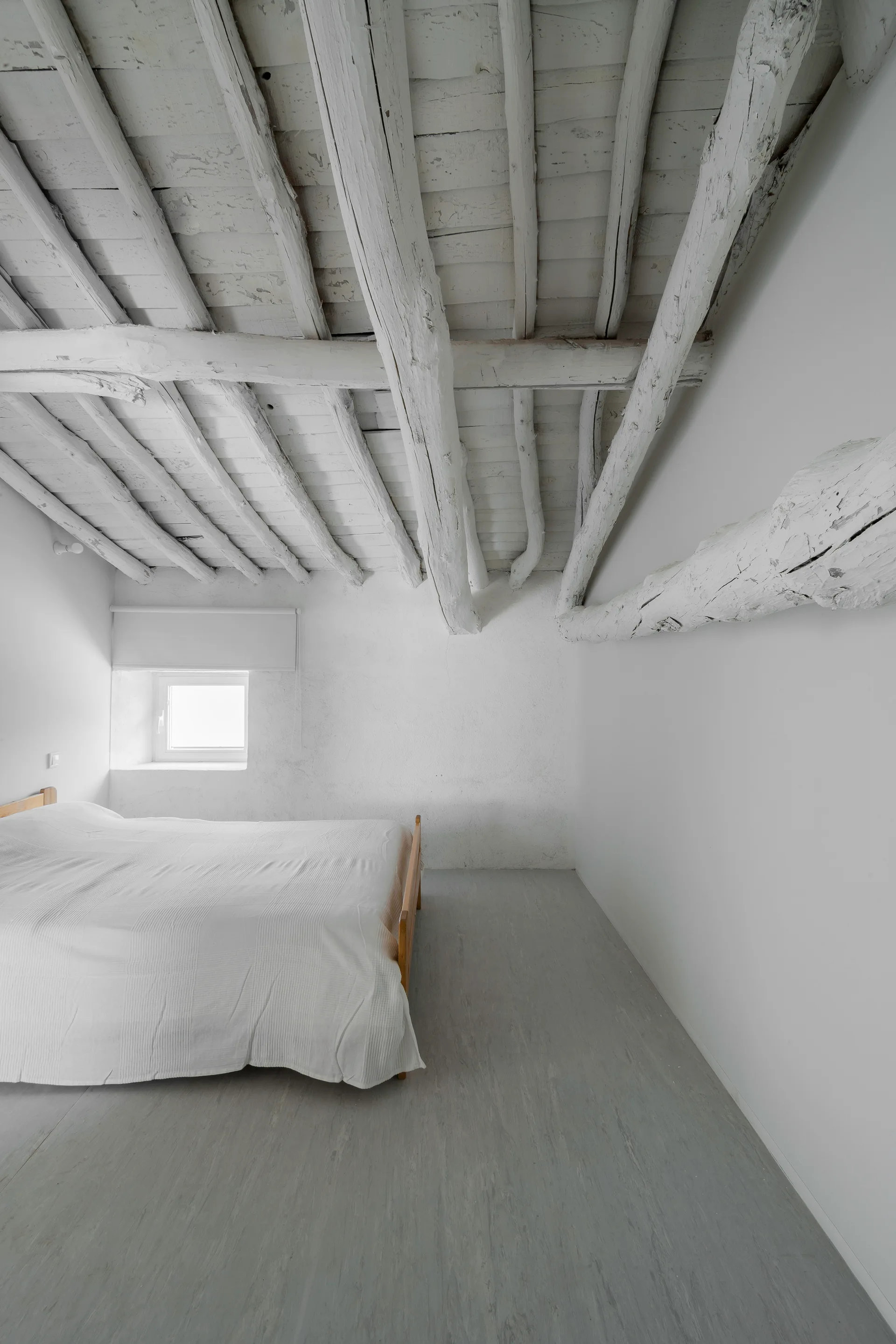A minimalist bedroom featuring a simple wooden bed, grey flooring, white walls, and a rustic white-painted wooden log ceiling.