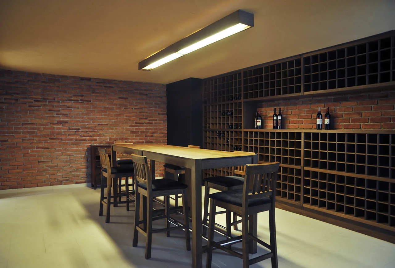 A wine cellar features a large wooden grid rack on an exposed brick wall next to a tall wood dining table with high-back stools.
