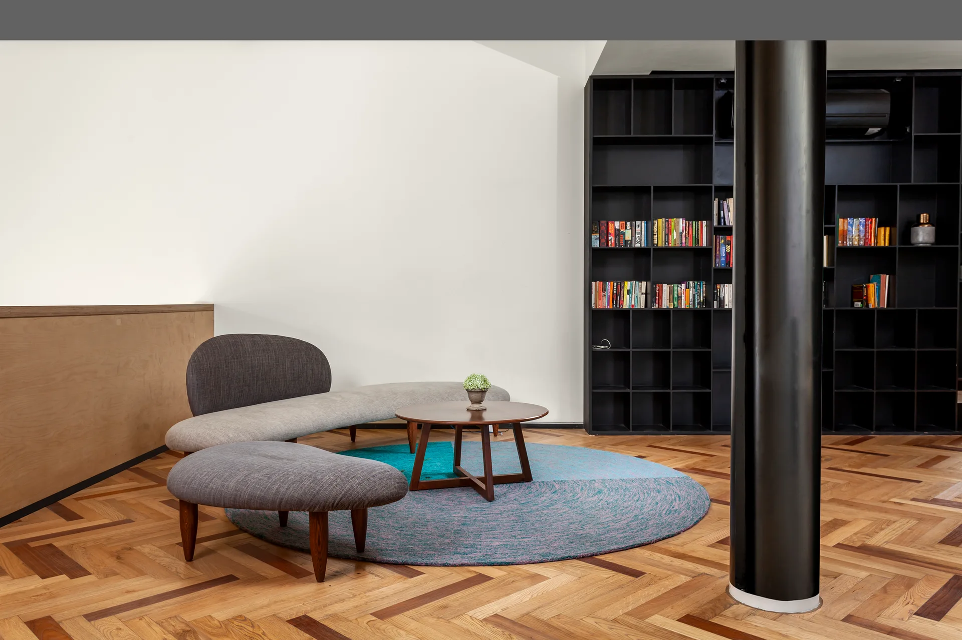 A modern living space features a curved grey sofa, a matching ottoman, a round rug, and a large black bookshelf on a herringbone wood floor.