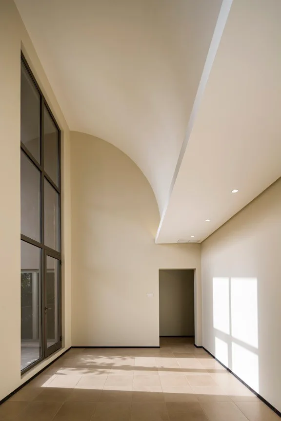 An empty minimalist room featuring a dramatic curved vaulted ceiling, tall multi-pane windows, and large-format beige floor tiles.