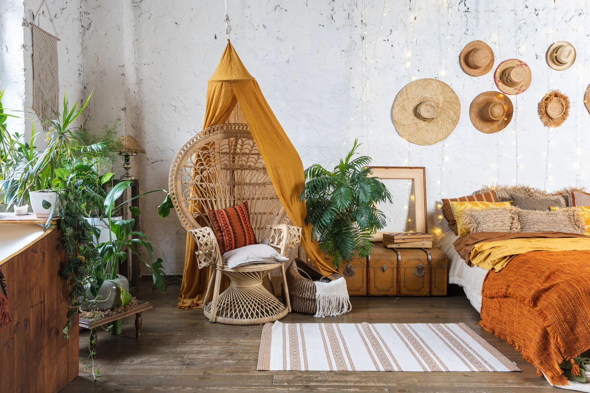 A wicker peacock chair sits beneath a mustard canopy next to a textured plaster wall decorated with straw hats in a bohemian bedroom.