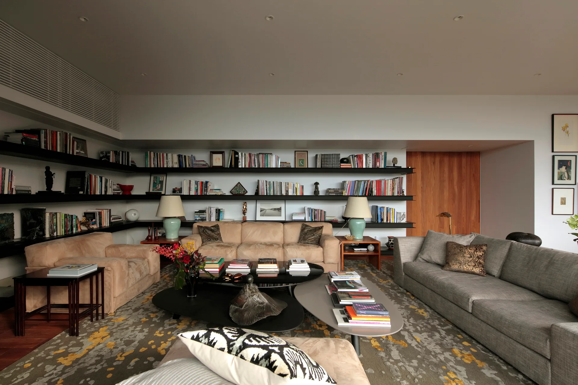 A modern living room features gray and beige sofas, nested coffee tables, and long black floating bookshelves filled with books along the wall.