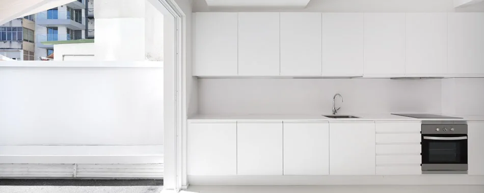 A minimalist all-white kitchen features flat-panel handleless cabinets, a stainless steel sink, an integrated oven, and a wide doorway opening to a balcony.
