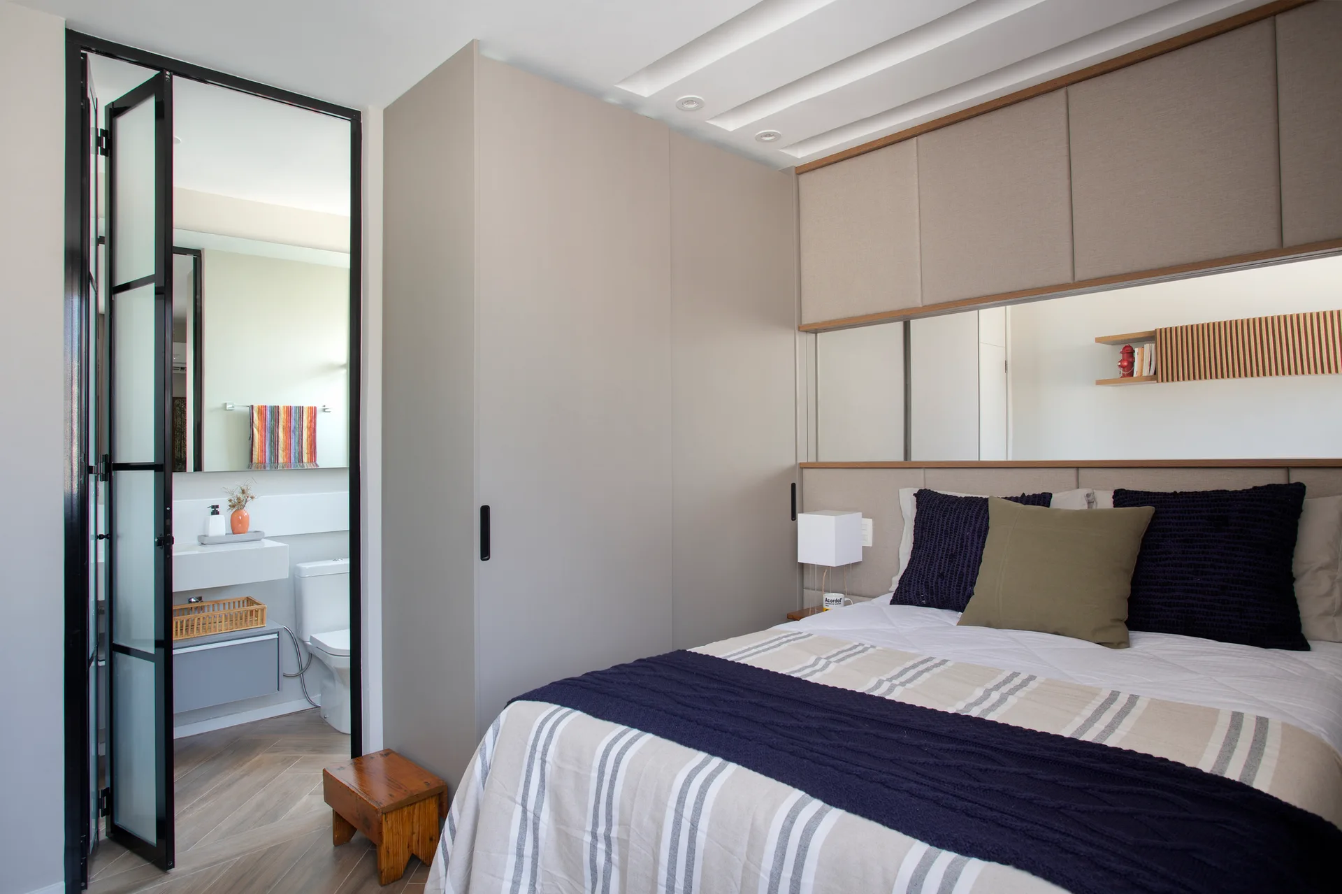 A modern bedroom featuring a built-in grey wardrobe, an upholstered headboard wall with a horizontal mirror, and a bathroom seen through a glass door.