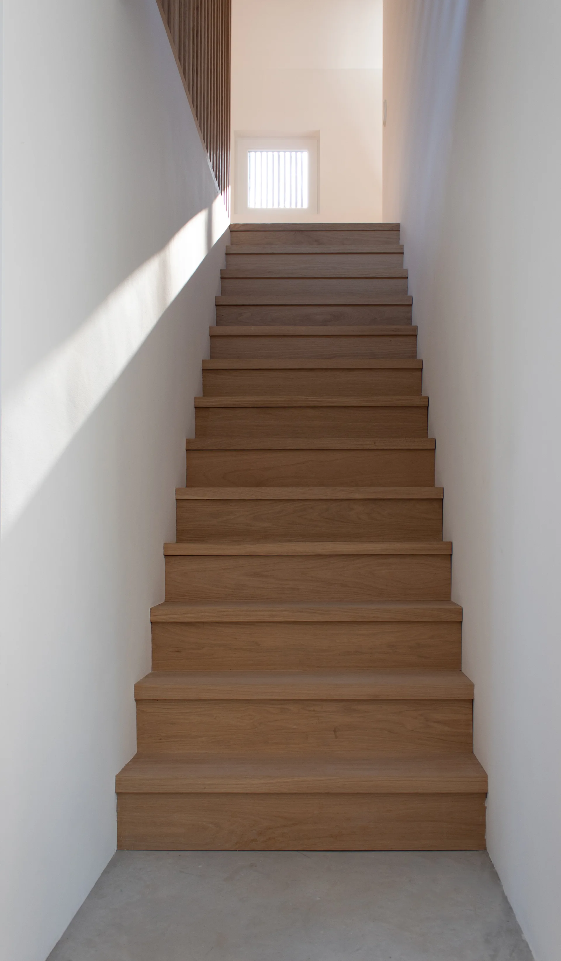 A straight-on view of a minimalist staircase with natural wood treads set between smooth white walls, leading up to a small window.