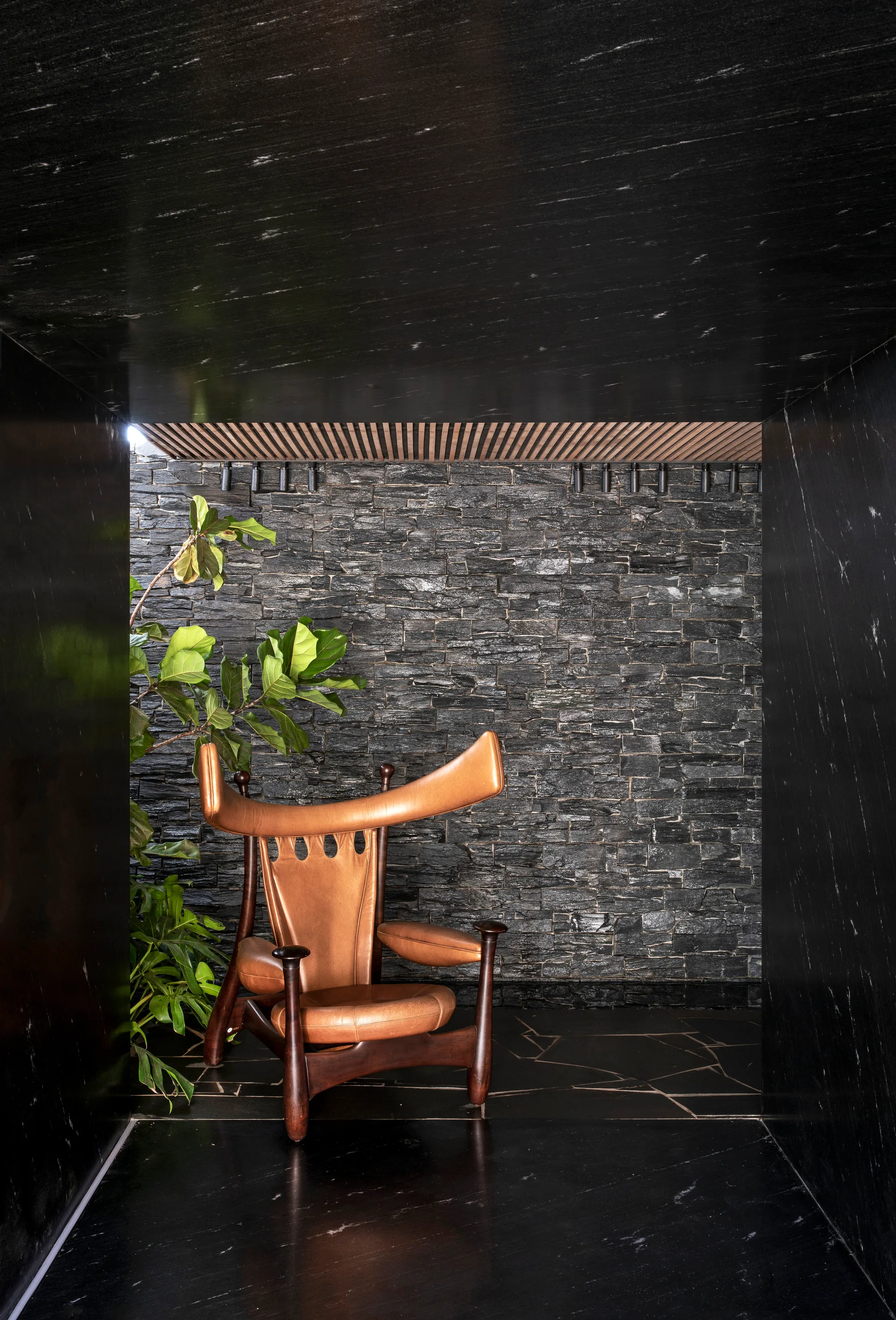 A sculptural tan leather armchair is positioned inside a dark alcove framed by polished black marble and backed by a textured stone wall.