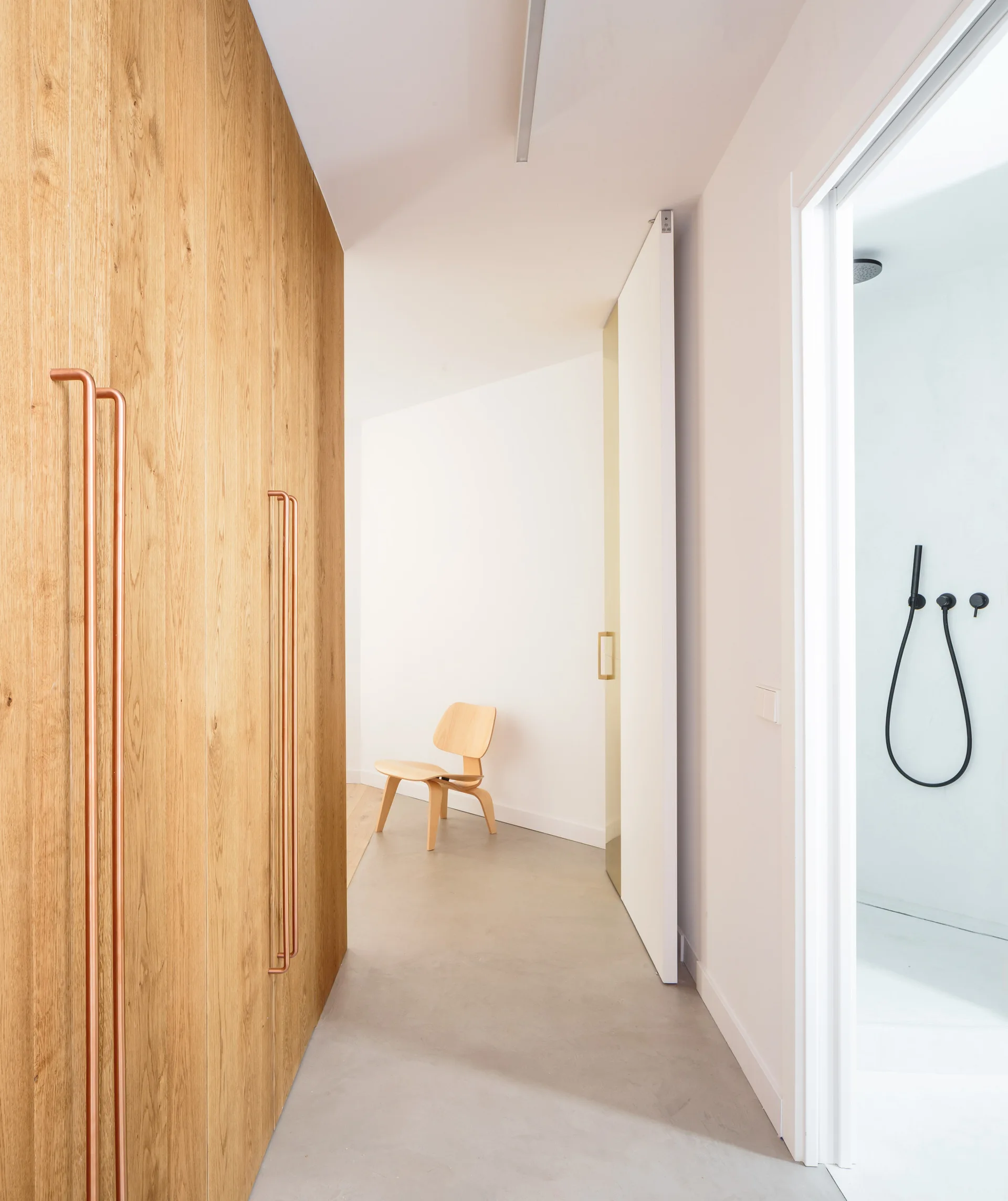 A minimalist hallway featuring floor-to-ceiling wood cabinets with long vertical copper handles next to a bright bathroom doorway.