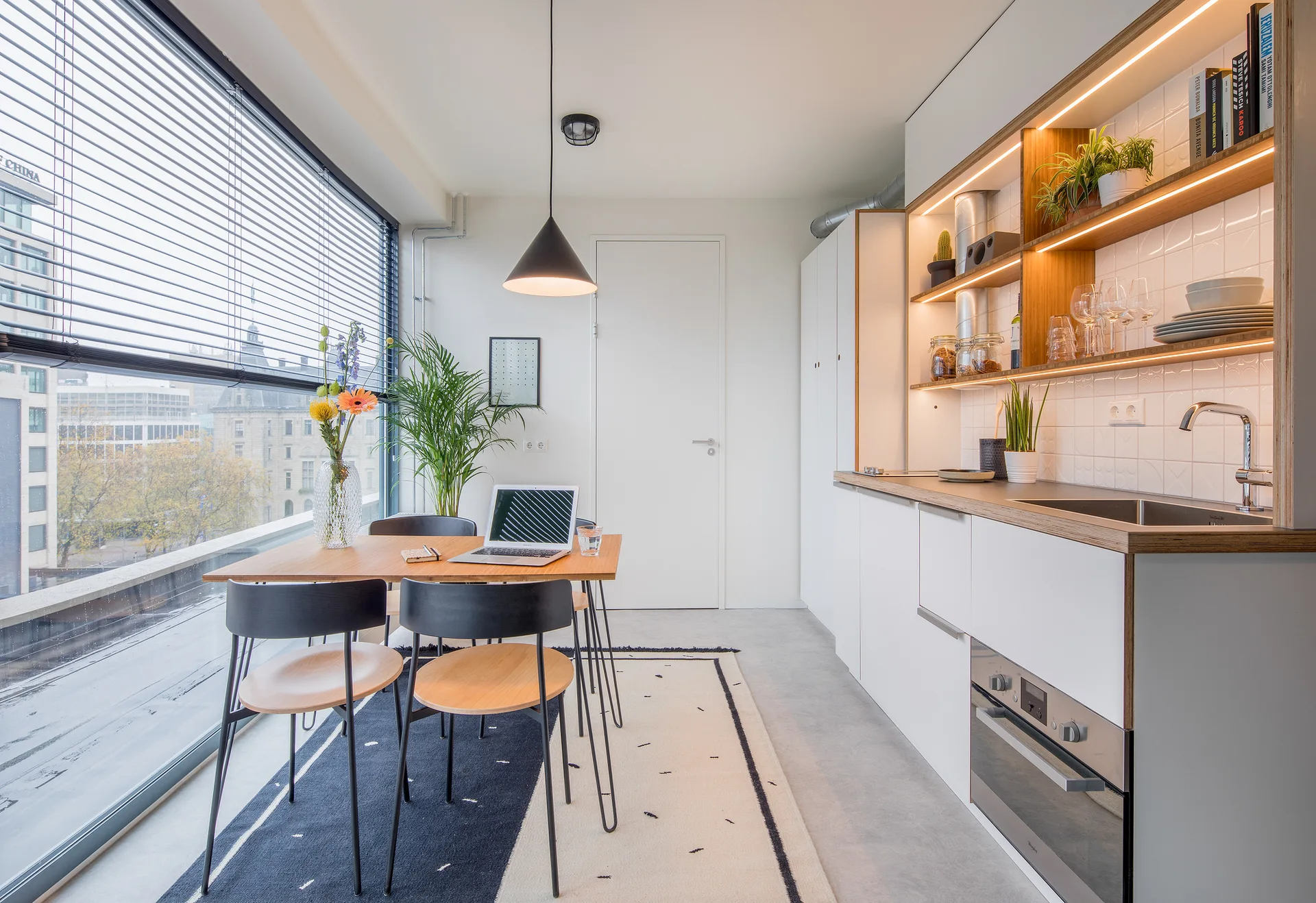 A modern apartment kitchen featuring white cabinetry, backlit open wooden shelves, and a wooden dining table with black chairs near a large window.