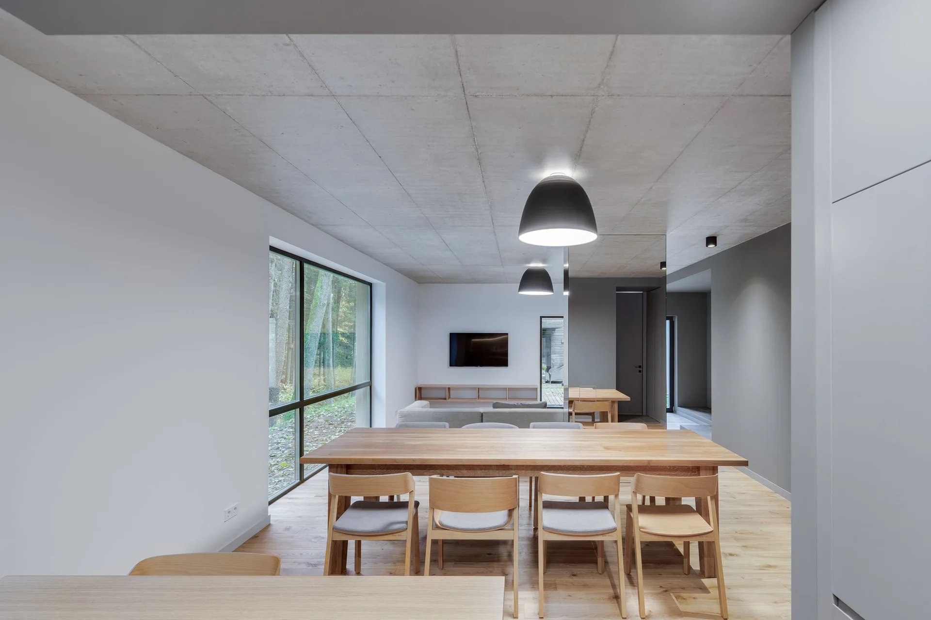 A modern dining area features a light wood table and chairs beneath an exposed concrete ceiling with a large window looking out onto trees.