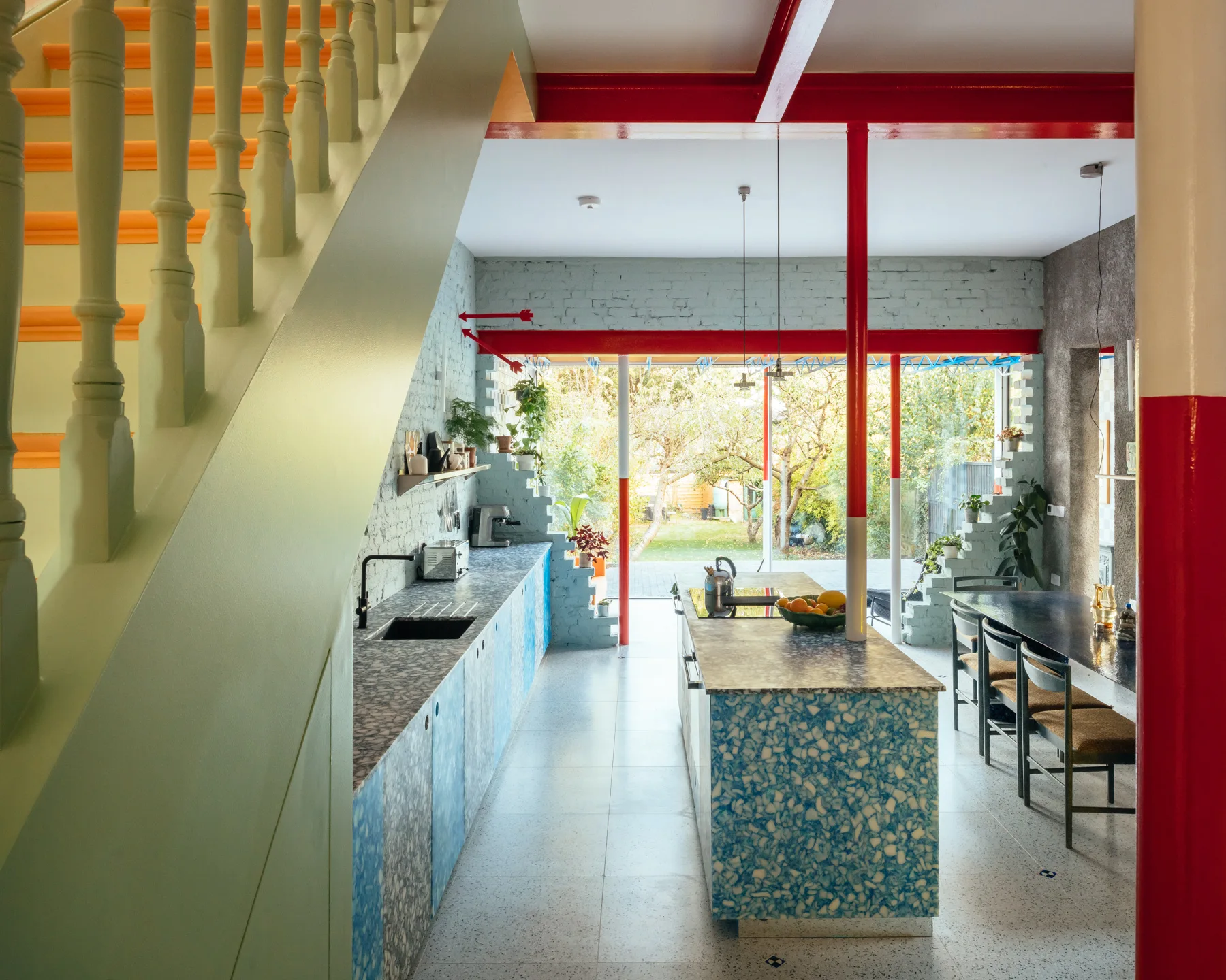 An open kitchen features a blue terrazzo-patterned island, a light green staircase, and bright red structural beams running across the white ceiling.