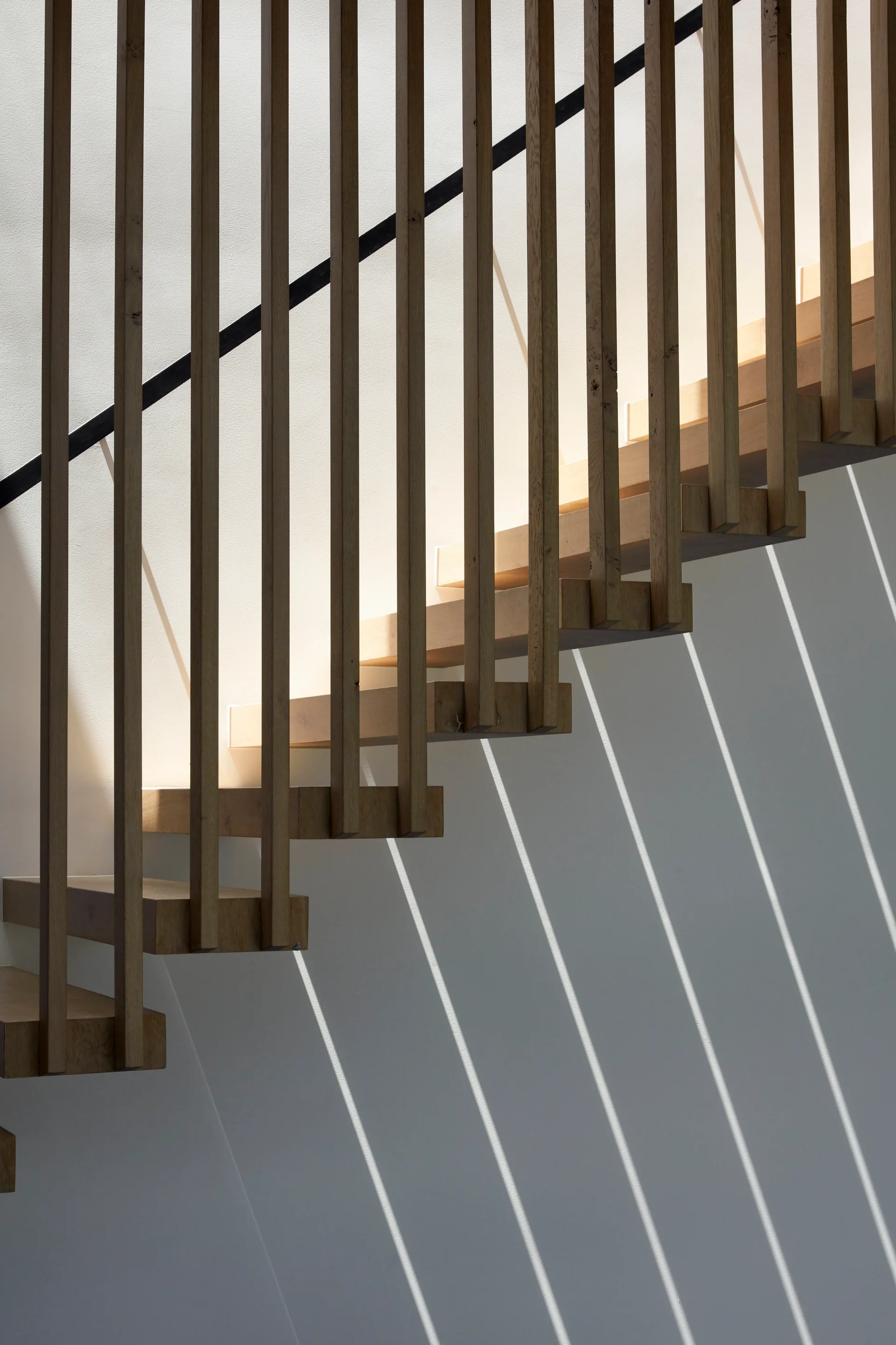 A minimalist wooden staircase with vertical slat balustrades and integrated step lighting casting diagonal shadows on a plain white wall.