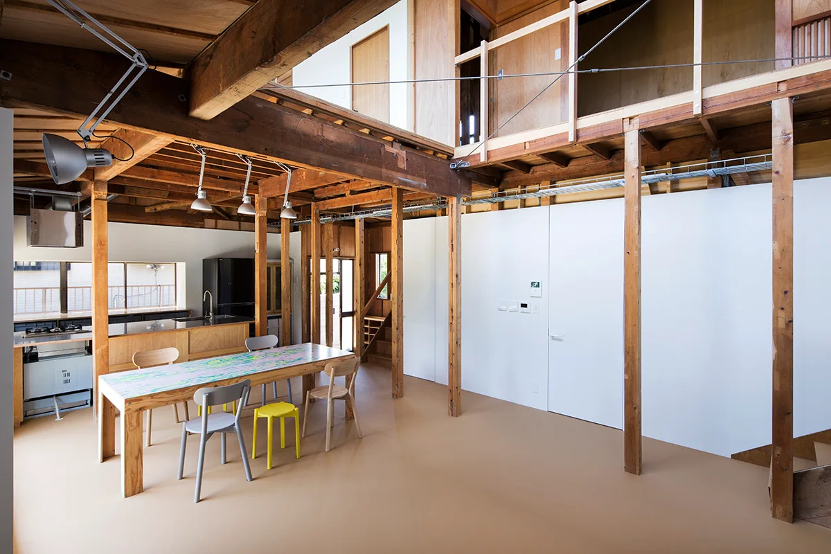 An open dining room features exposed wood structural columns and beams, a tan floor, a long patterned table, and a white partition wall.