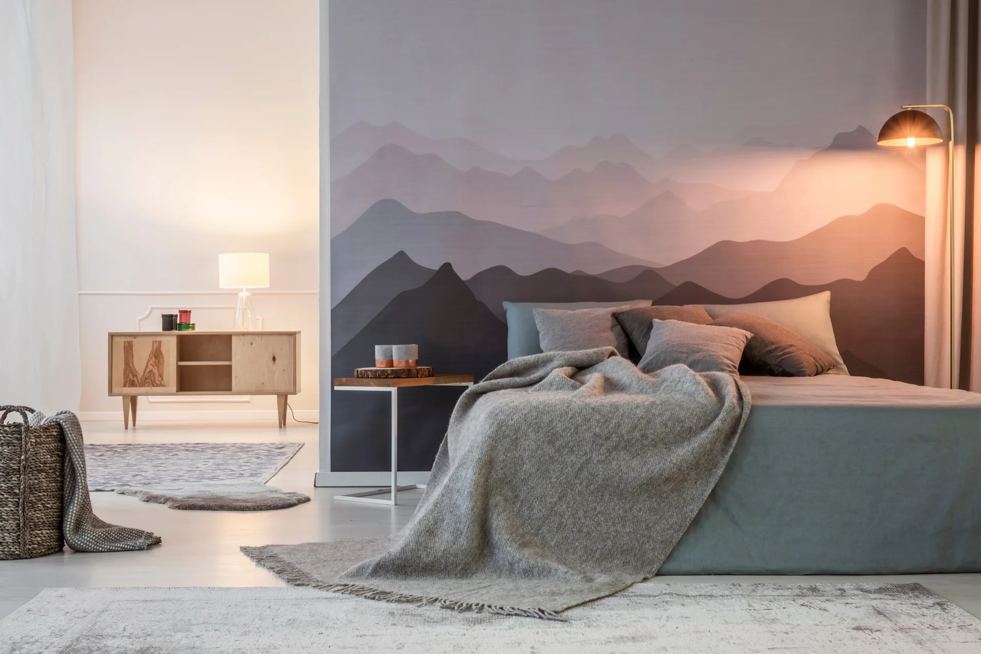 A gray-toned bed with a soft throw sits against a wall decorated with a purple mountain landscape mural next to a gold floor lamp.