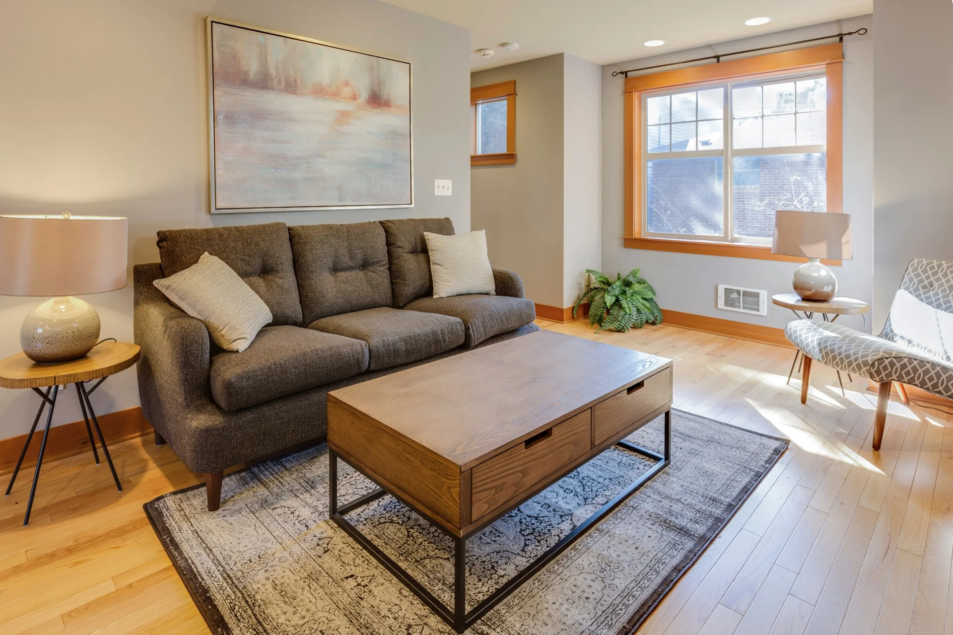 A gray living room features a dark charcoal sofa, a wooden coffee table over a patterned rug, and a large window with warm wood trim.