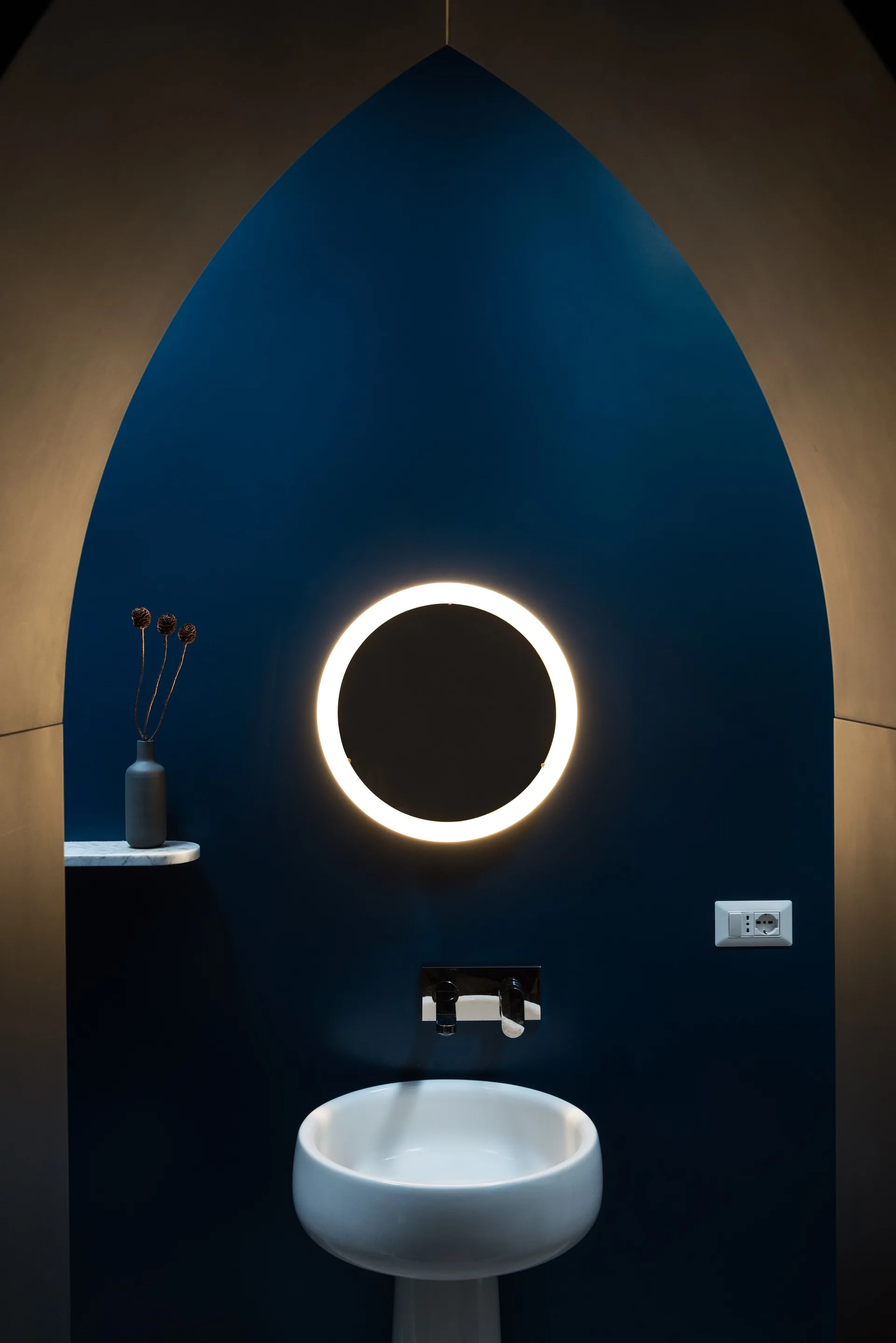 A white pedestal bathroom sink and faucet set against a deep blue arched alcove with a glowing round backlit mirror and a small marble shelf.