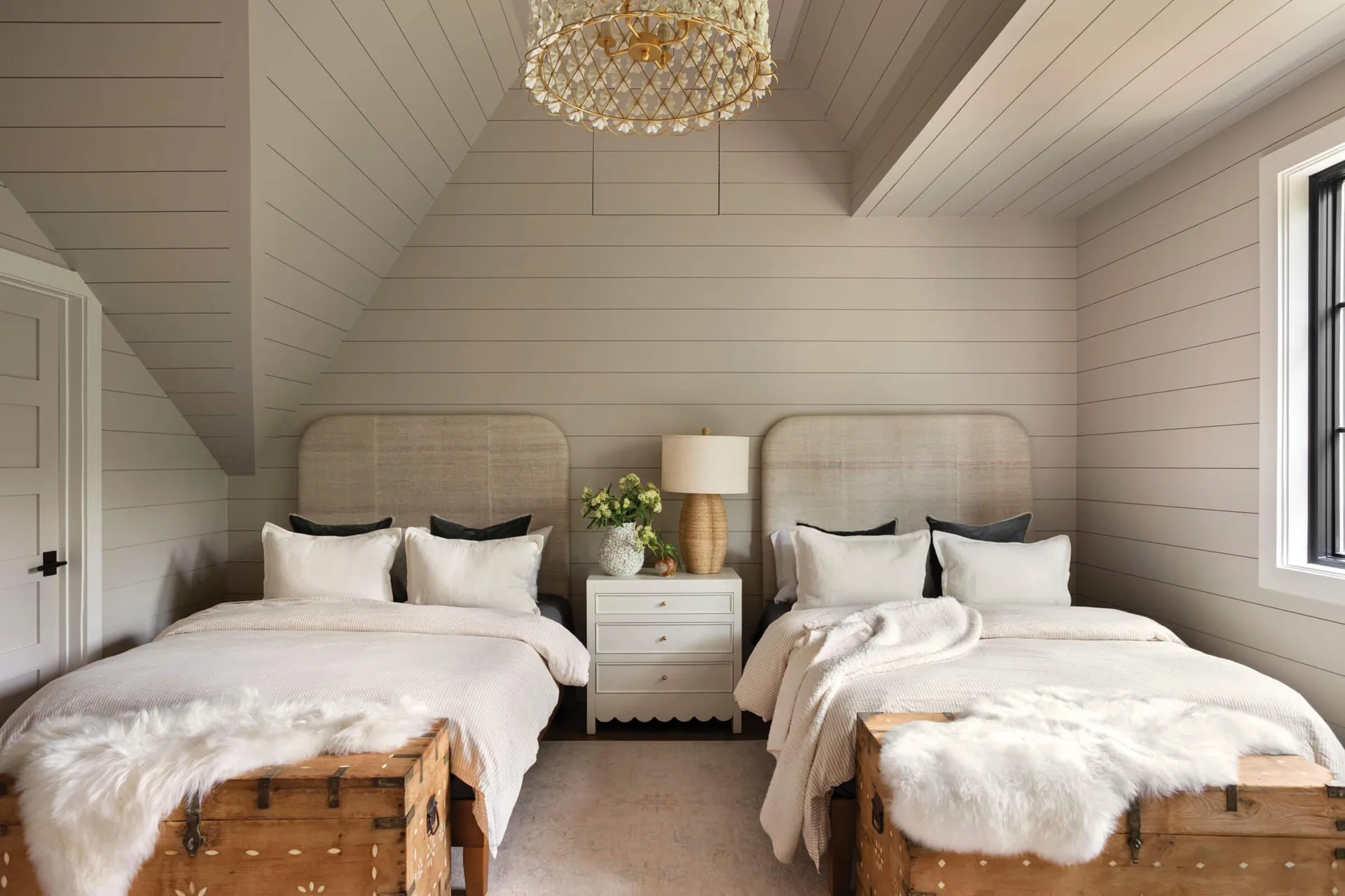Two twin beds with upholstered headboards stand symmetrically against a beige shiplap wall, separated by a white nightstand.