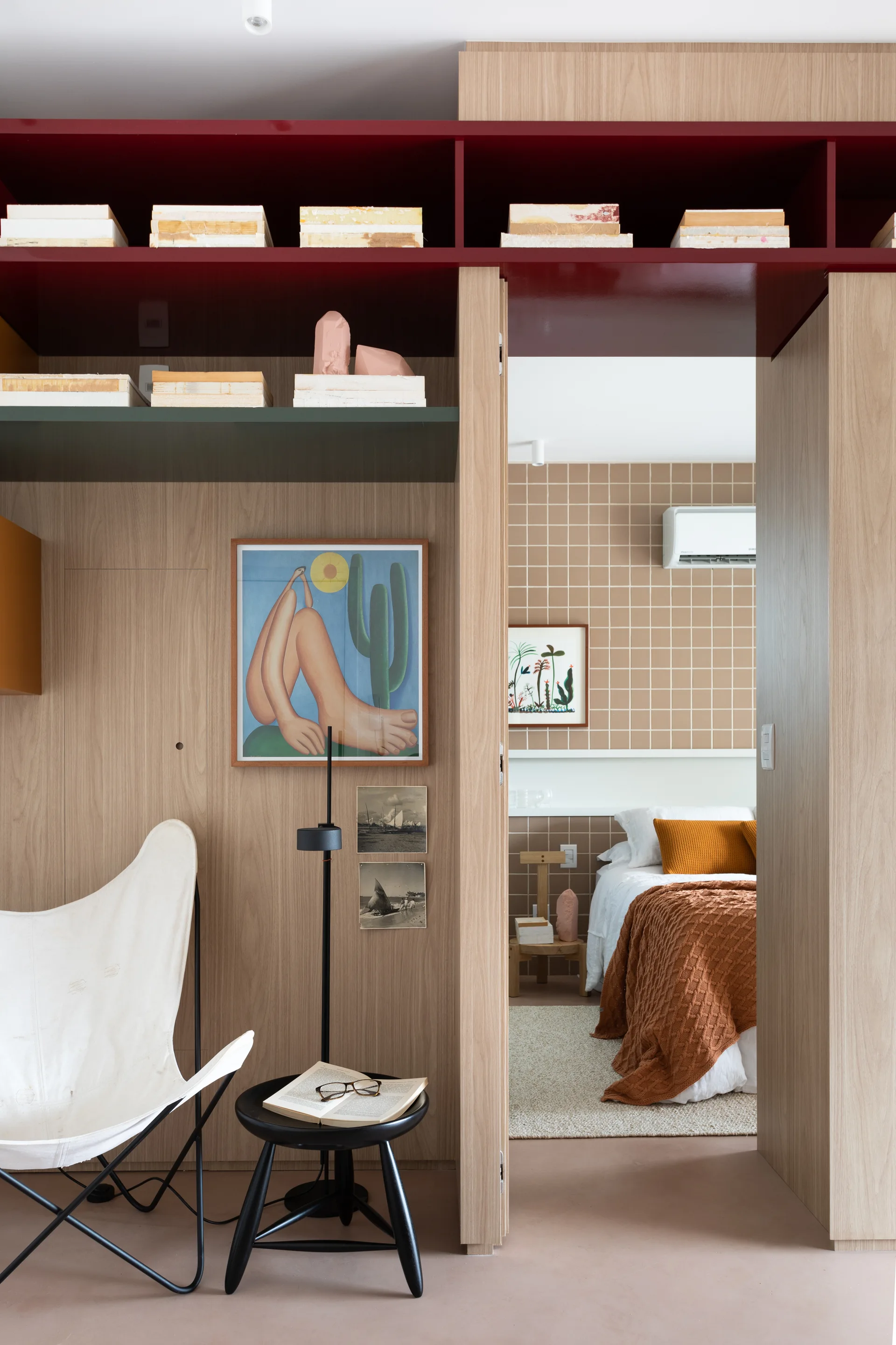 A view through a wood-paneled doorway reveals a bedroom with a grid-tiled wall, while a butterfly chair and red shelf sit in the foreground.