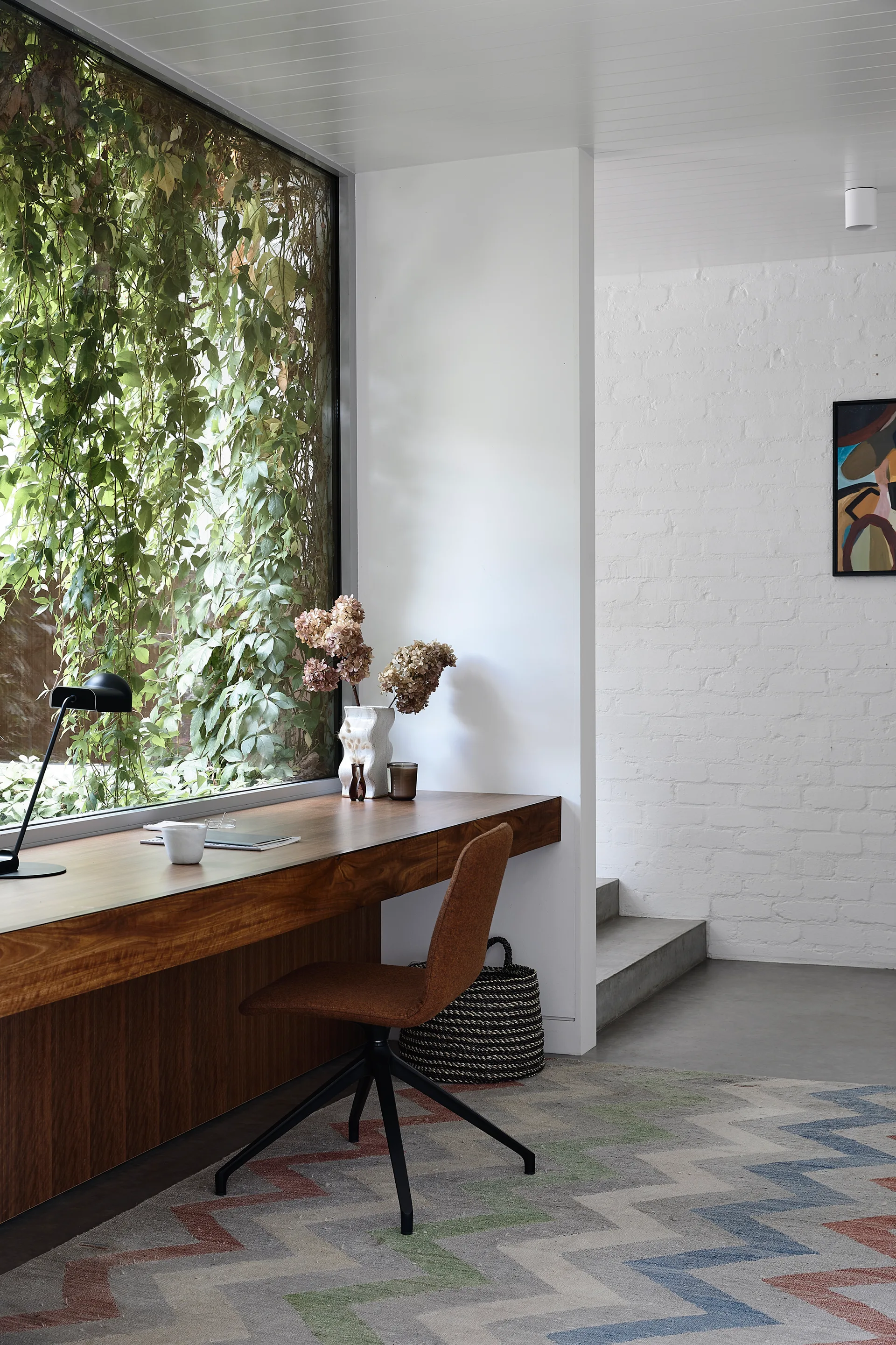 A long wooden desk sits in front of a large window filled with green ivy next to a white painted brick wall and a brown office chair.