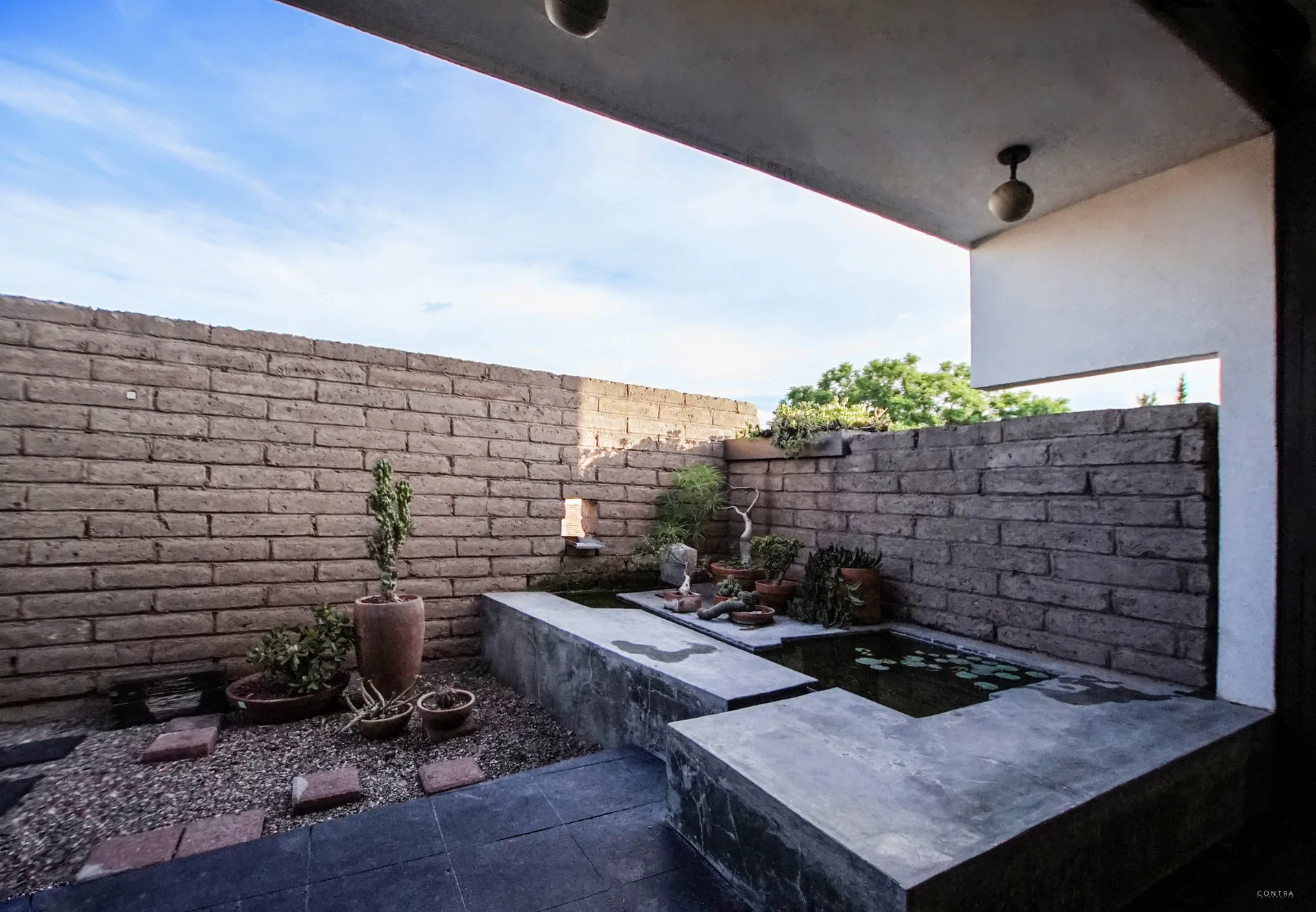 A minimalist outdoor courtyard features a concrete built-in pond with water lilies, surrounded by rough brick walls and potted plants.