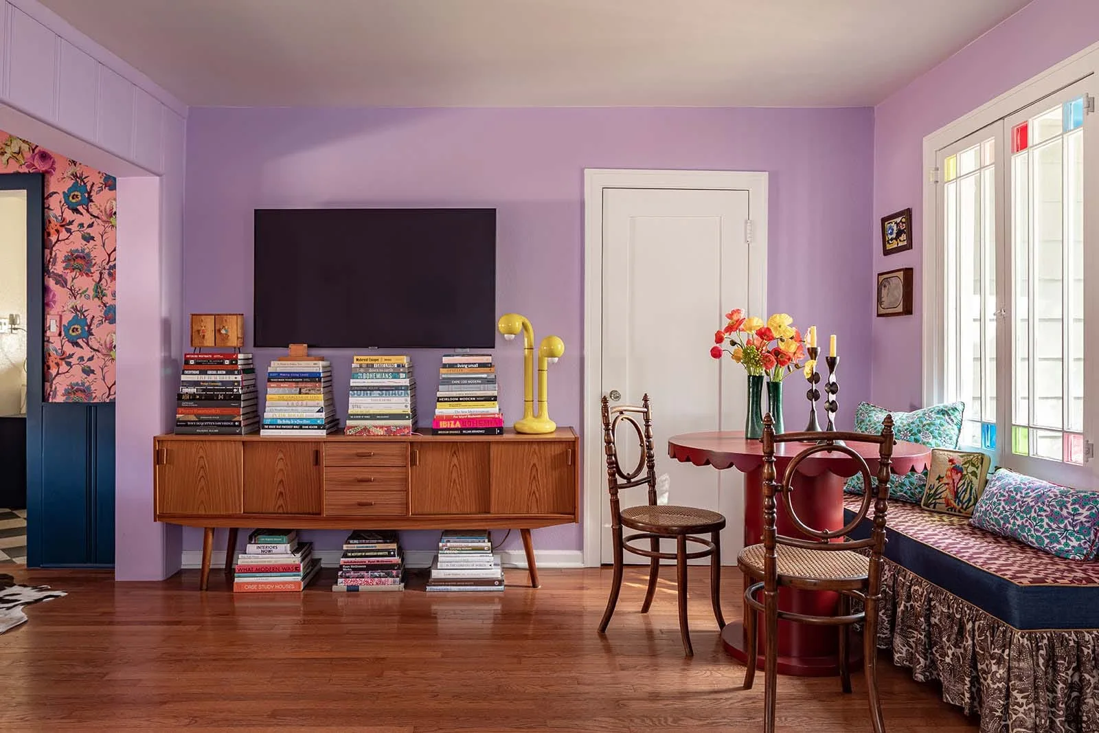 An eclectic dining room features lavender walls, a wood credenza topped with book stacks, and a red scalloped table beside a patterned window seat.