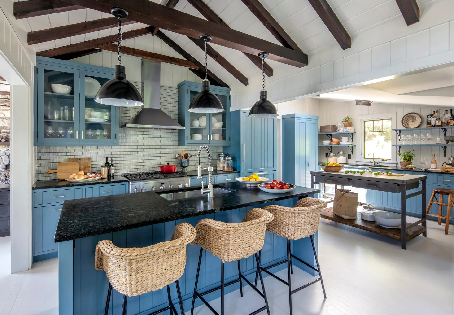 A kitchen with blue cabinets, a black-topped island with three wicker bar stools, grey subway tile backsplash, and dark exposed ceiling beams.