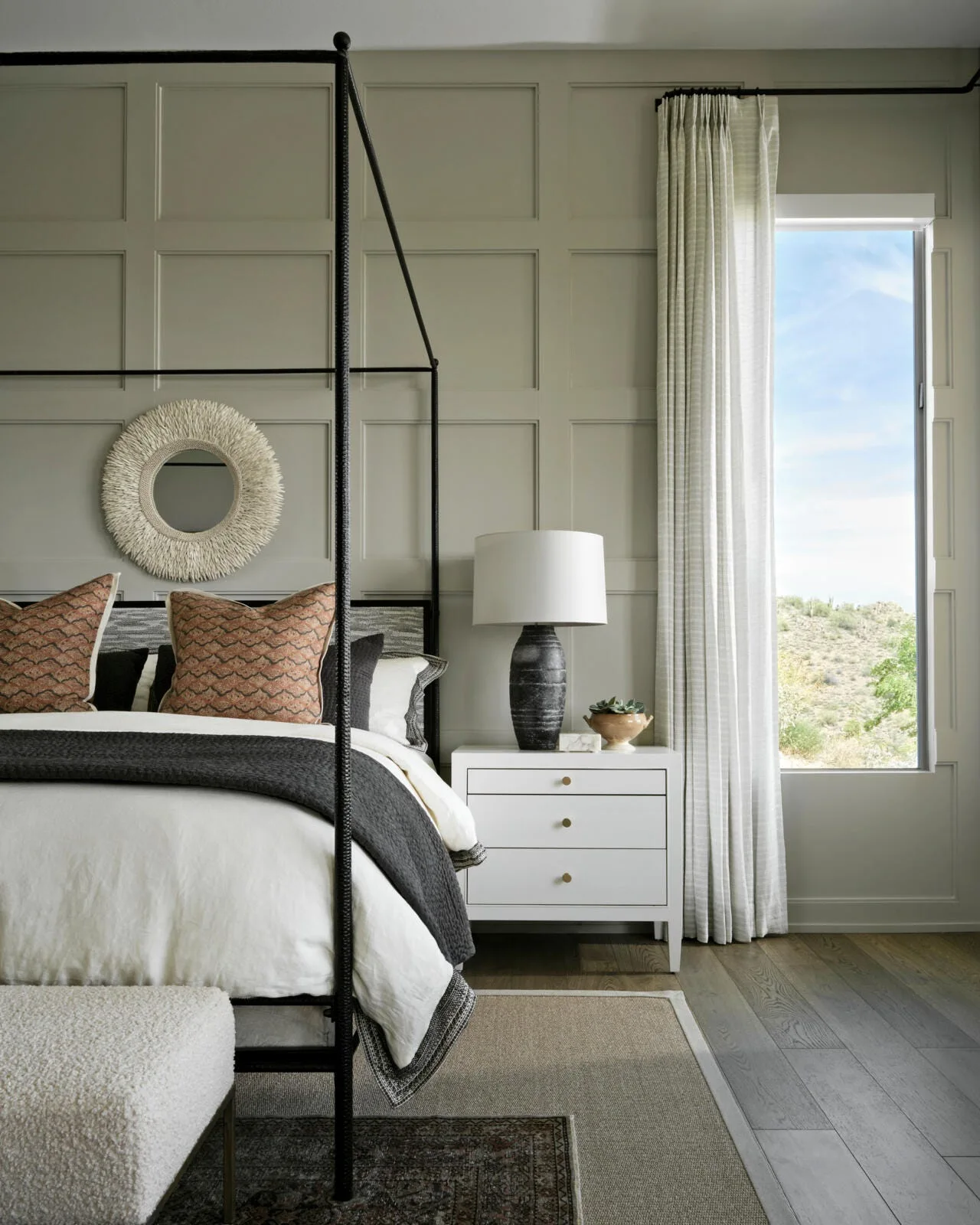 A modern bedroom features a black metal canopy bed against a sage green paneled wall next to a white nightstand and a tall window.