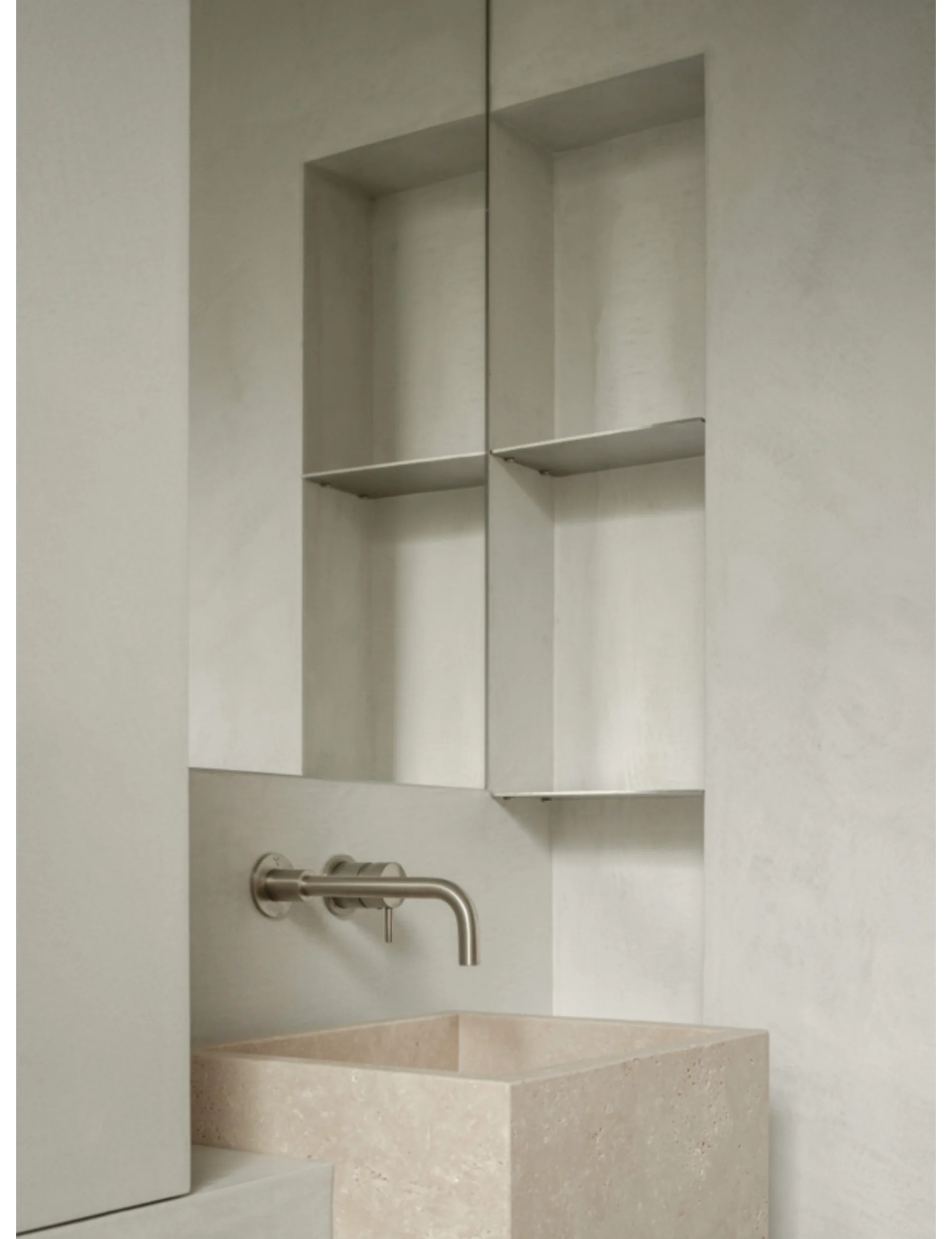 A minimalist bathroom features a square textured stone washbasin, a wall-mounted stainless steel faucet, and a built-in plaster wall niche.