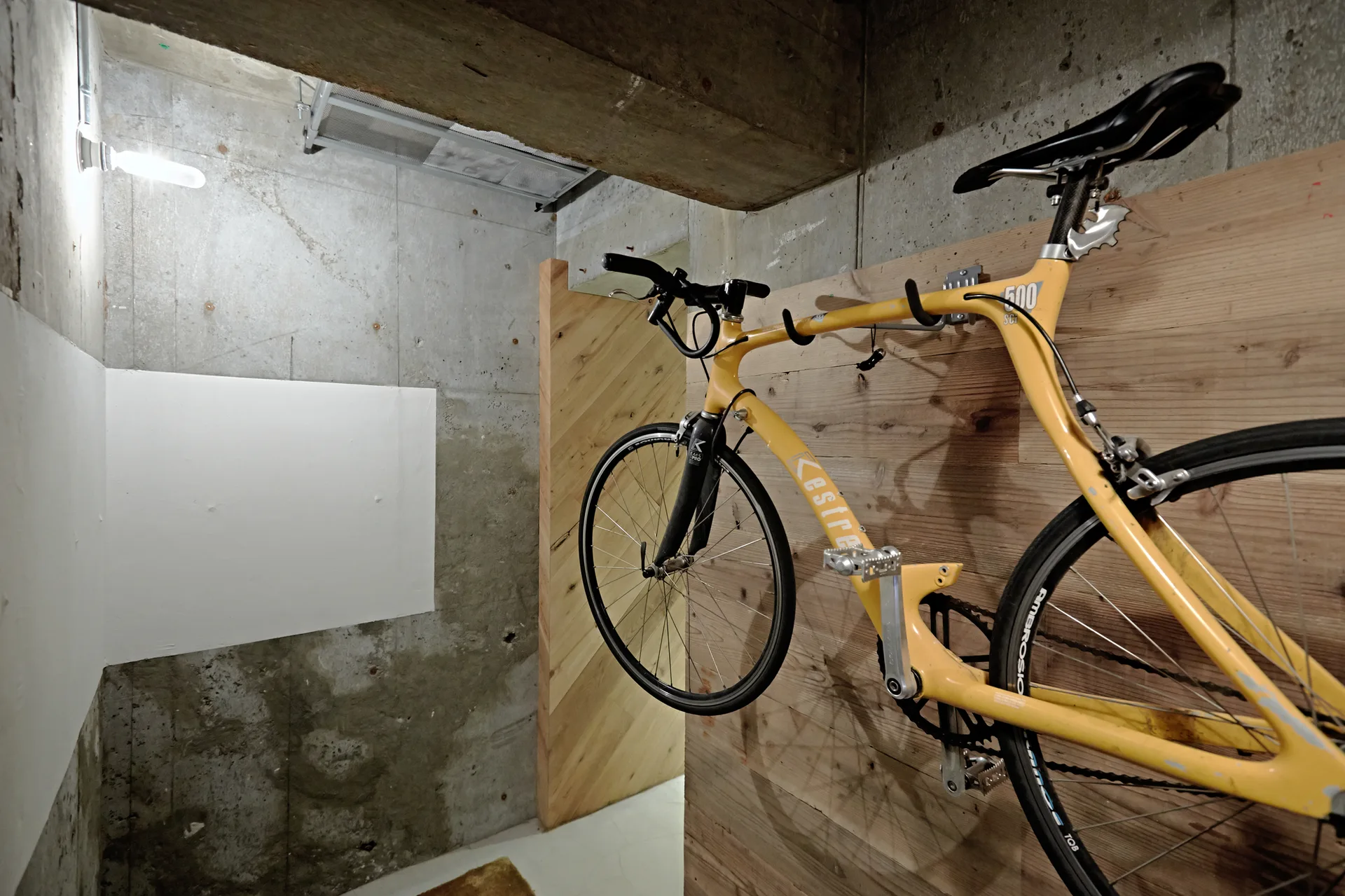 A yellow road bicycle is mounted horizontally on a light wood paneled wall in an industrial room with raw concrete walls.