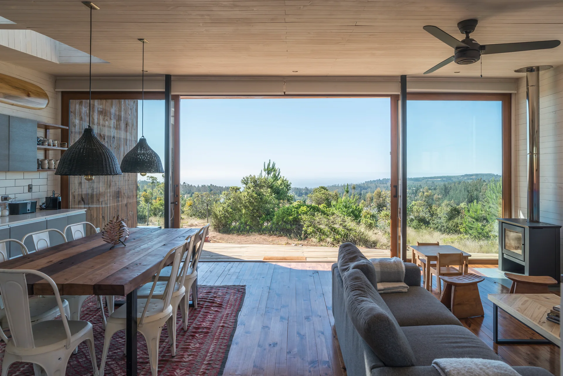 An open-concept cabin living and dining room features a large wood table, white metal chairs, and a grey sofa facing expansive sliding glass doors.