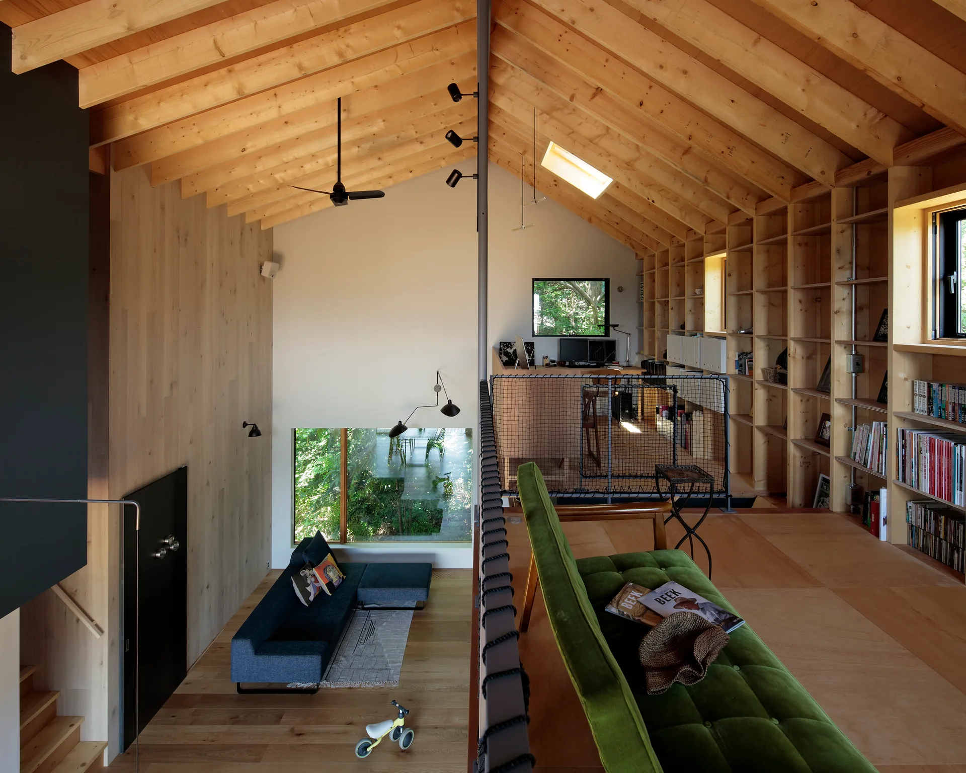 A split-level home interior with a lower lounge area, an upper mezzanine workspace, and extensive light wood built-in bookshelves.