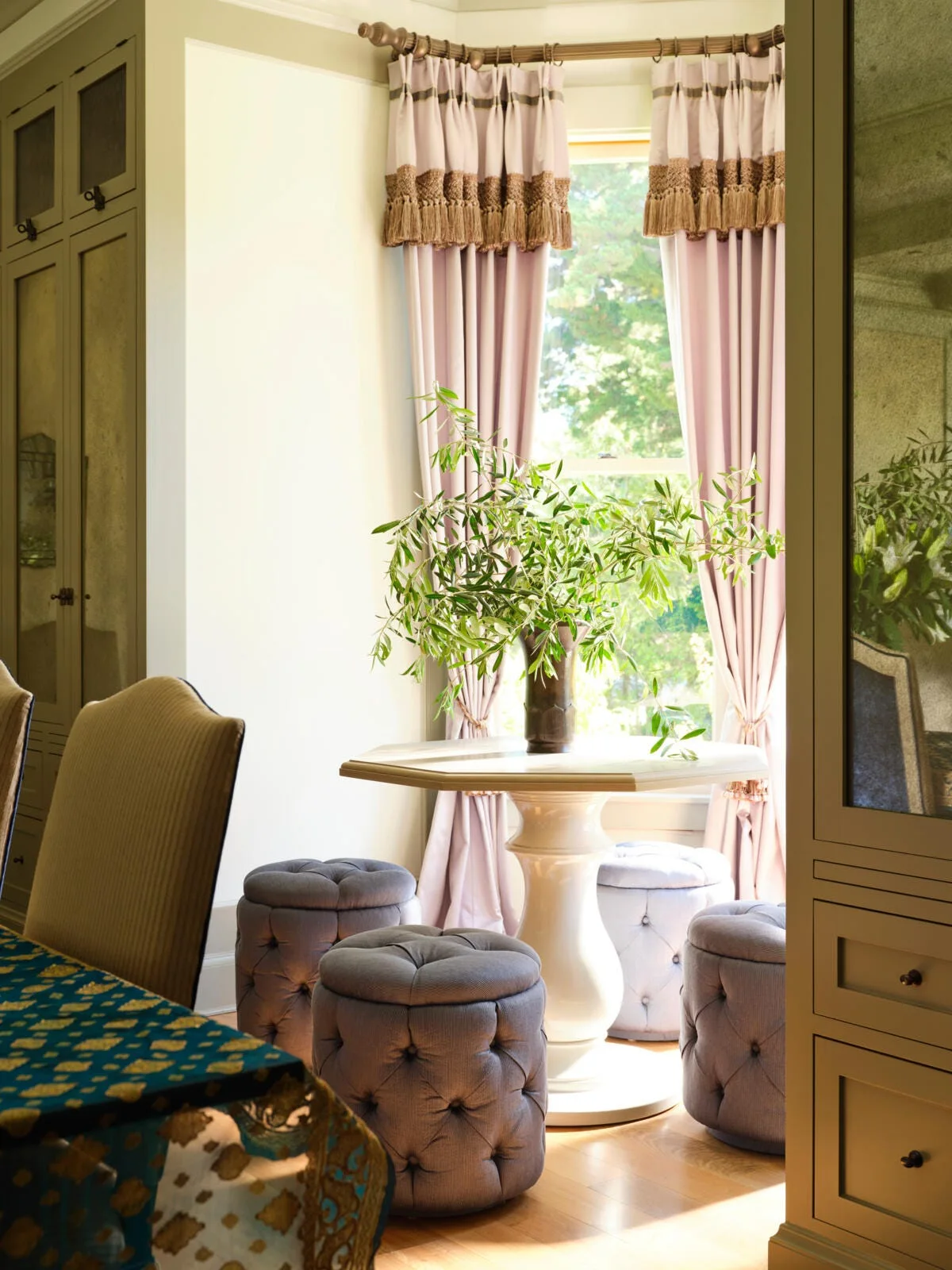 A white pedestal table is surrounded by tufted blue stools in a sunlit breakfast nook framed by lavender curtains and sage green cabinets.