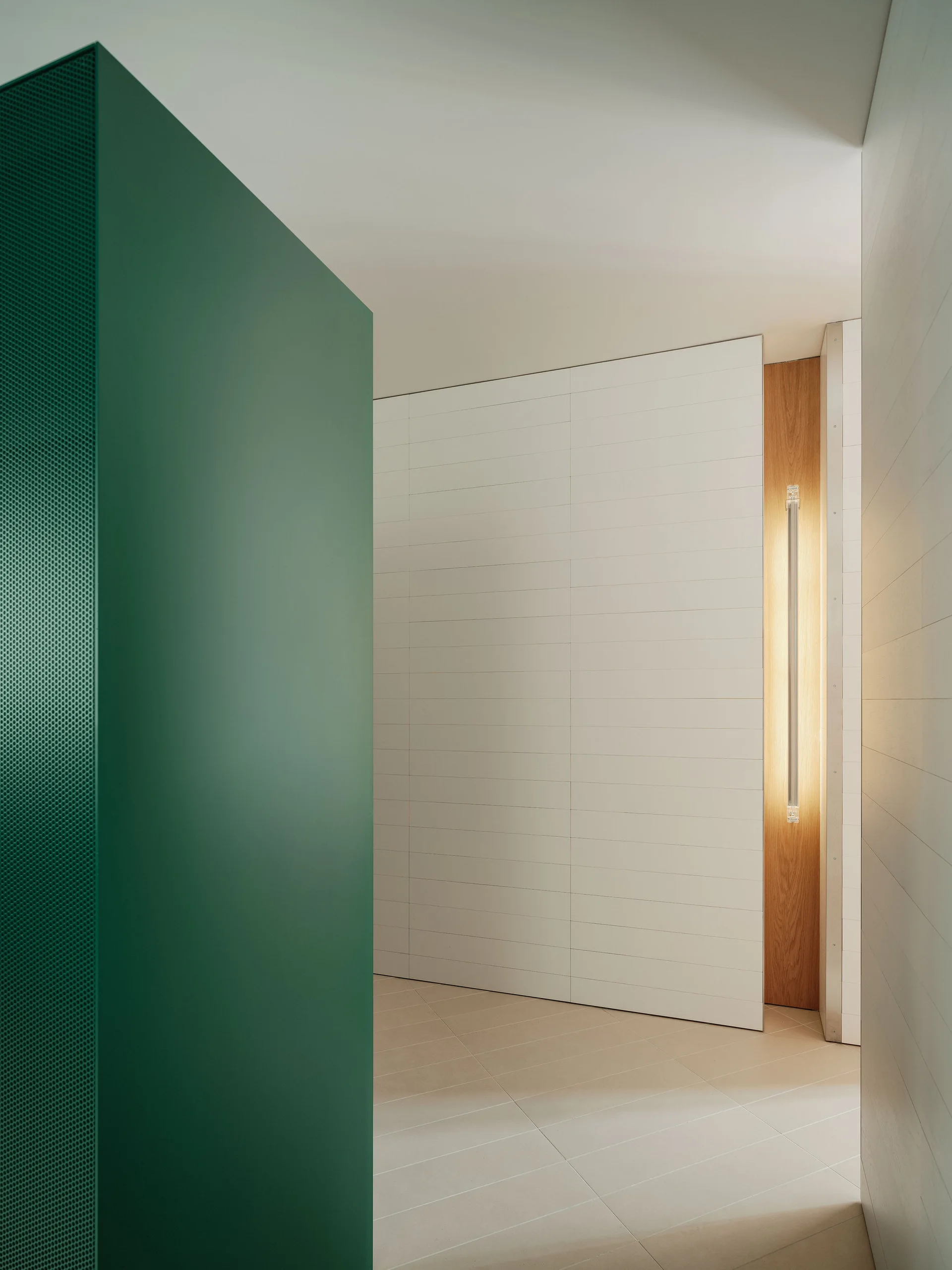 A minimalist hallway features a green perforated partition, white paneled walls, and a recessed wood-toned niche with a vertical integrated light.