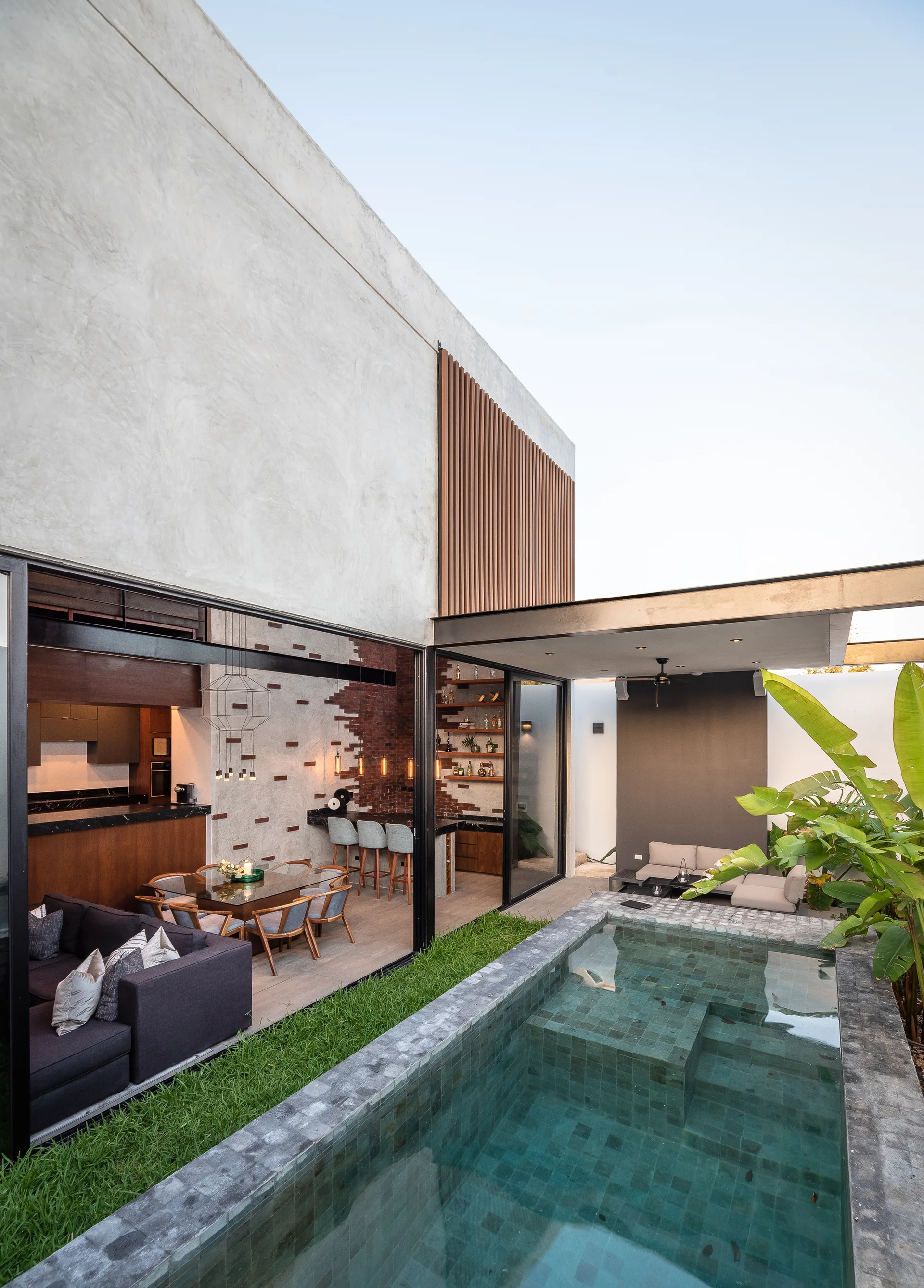 A modern concrete home features a swimming pool adjacent to sliding glass doors that open to a dining area.