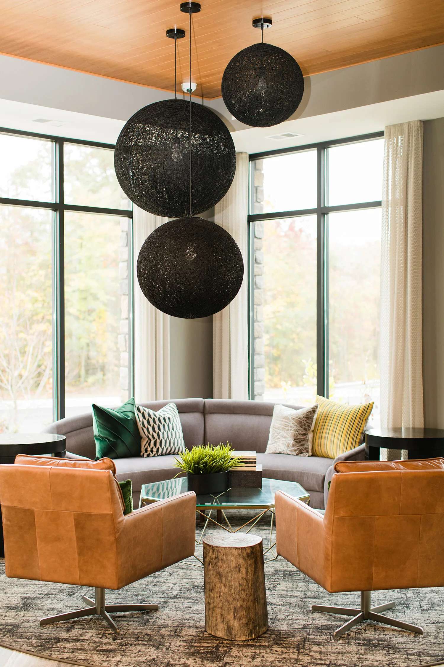 A modern seating area with a curved grey sofa, tan leather armchairs, a hexagonal glass coffee table, and large black woven sphere pendant lights.