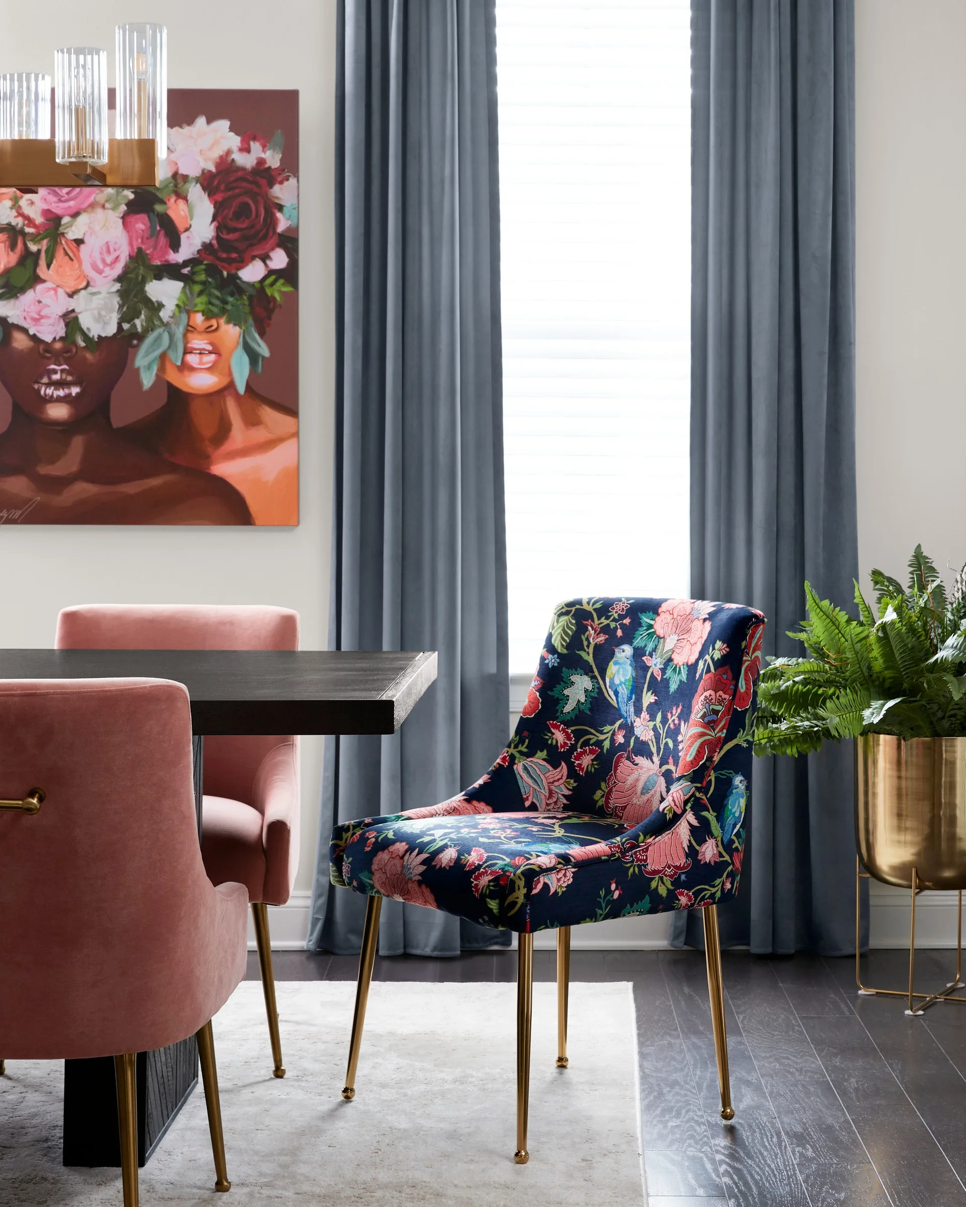 A dining room features a dark wood table with blush pink velvet chairs, a bold floral upholstered chair, blue curtains, and a gold fern planter.
