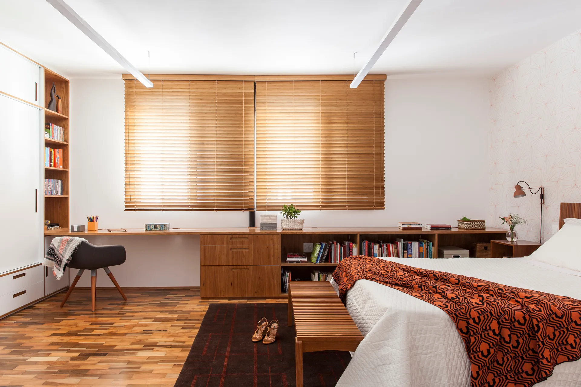 A modern bedroom featuring a low-profile wooden desk and shelving unit under wooden blinds next to a bed with an orange geometric throw blanket.