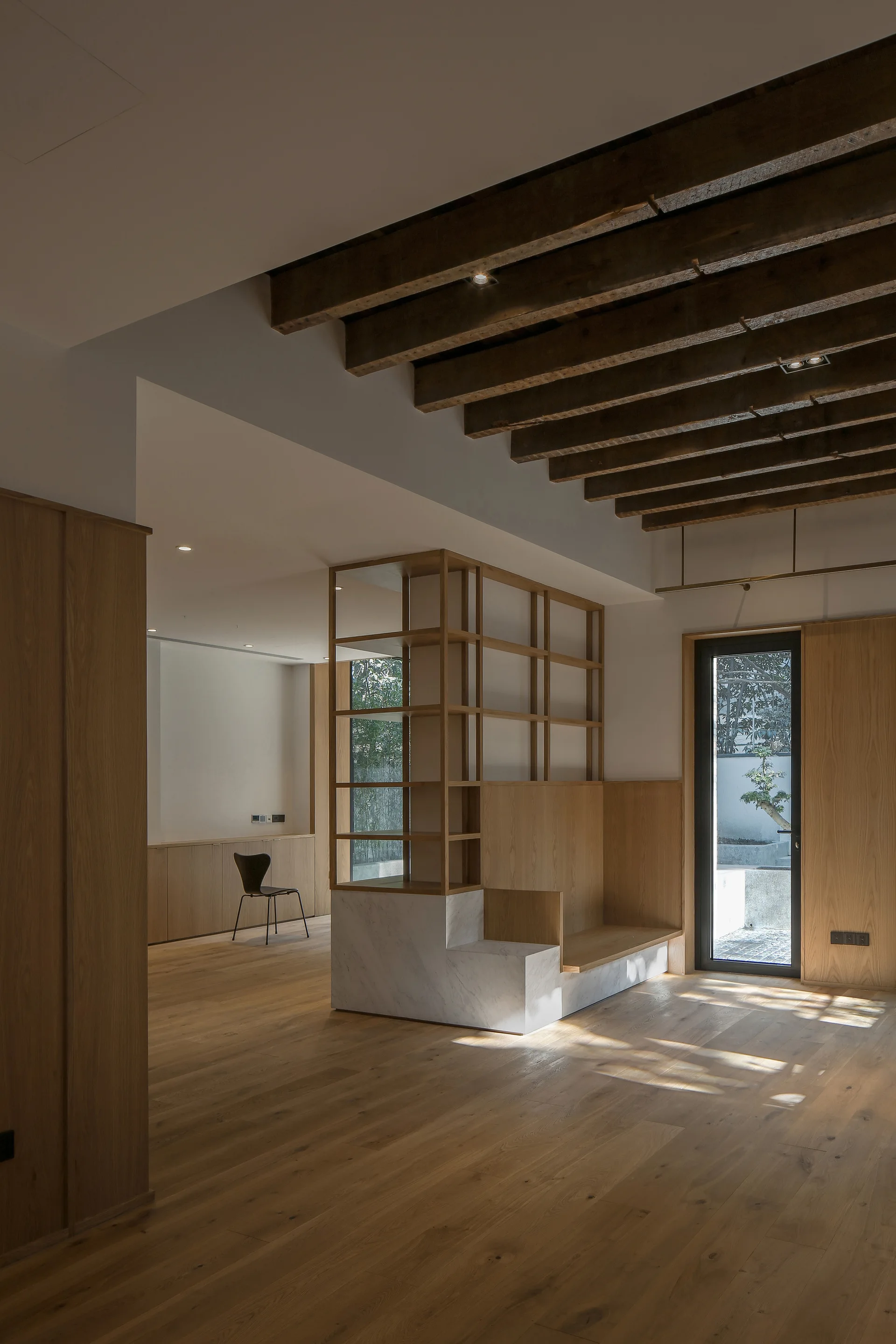 A minimalist living room features light wood flooring, exposed dark wooden ceiling beams, and a central partition unit with a white marble base.